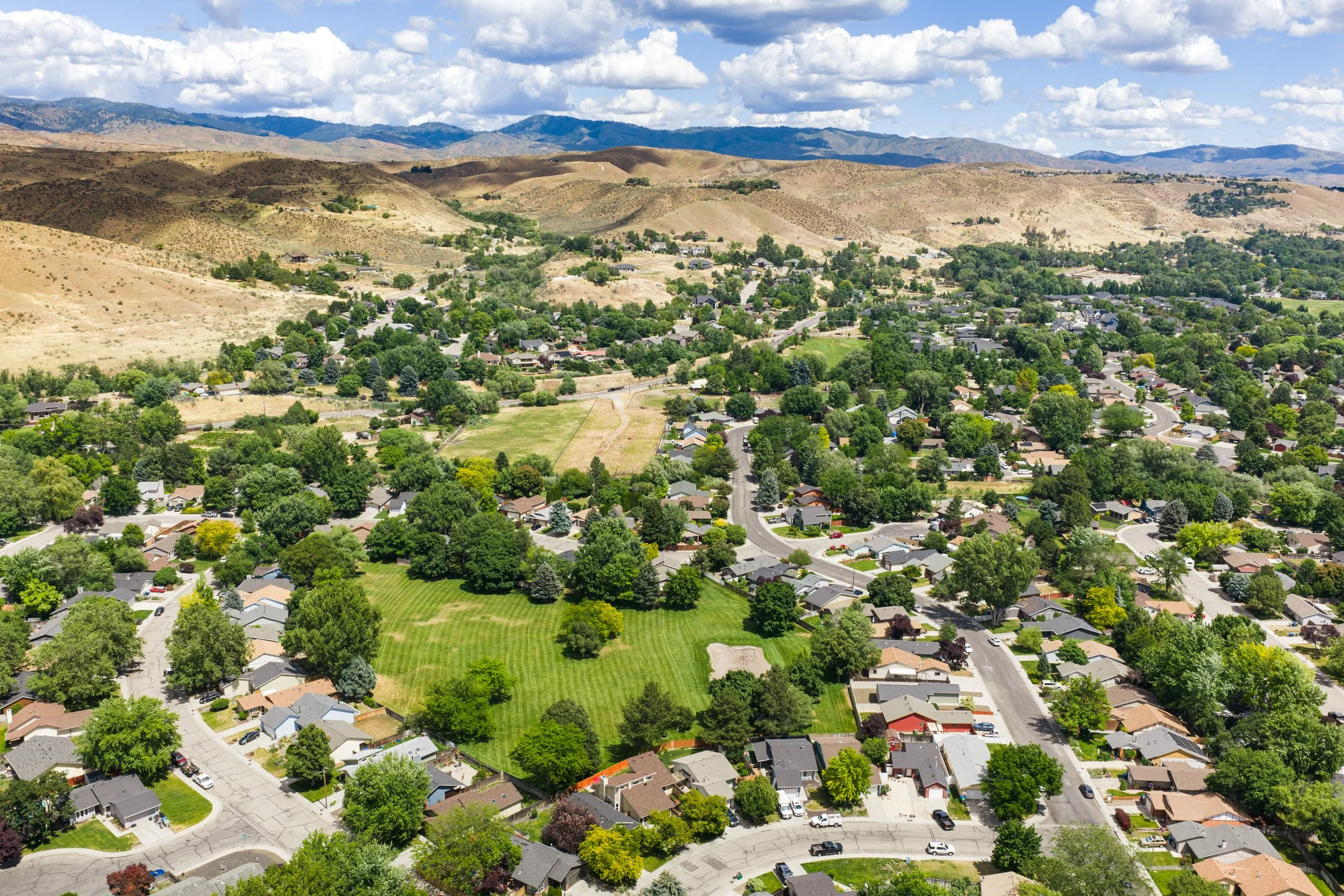 Aerial view of a residential neighborhood with houses, trees, green lawns, and winding streets, surrounded by rolling hills and mountainous terrain under a partly cloudy sky.