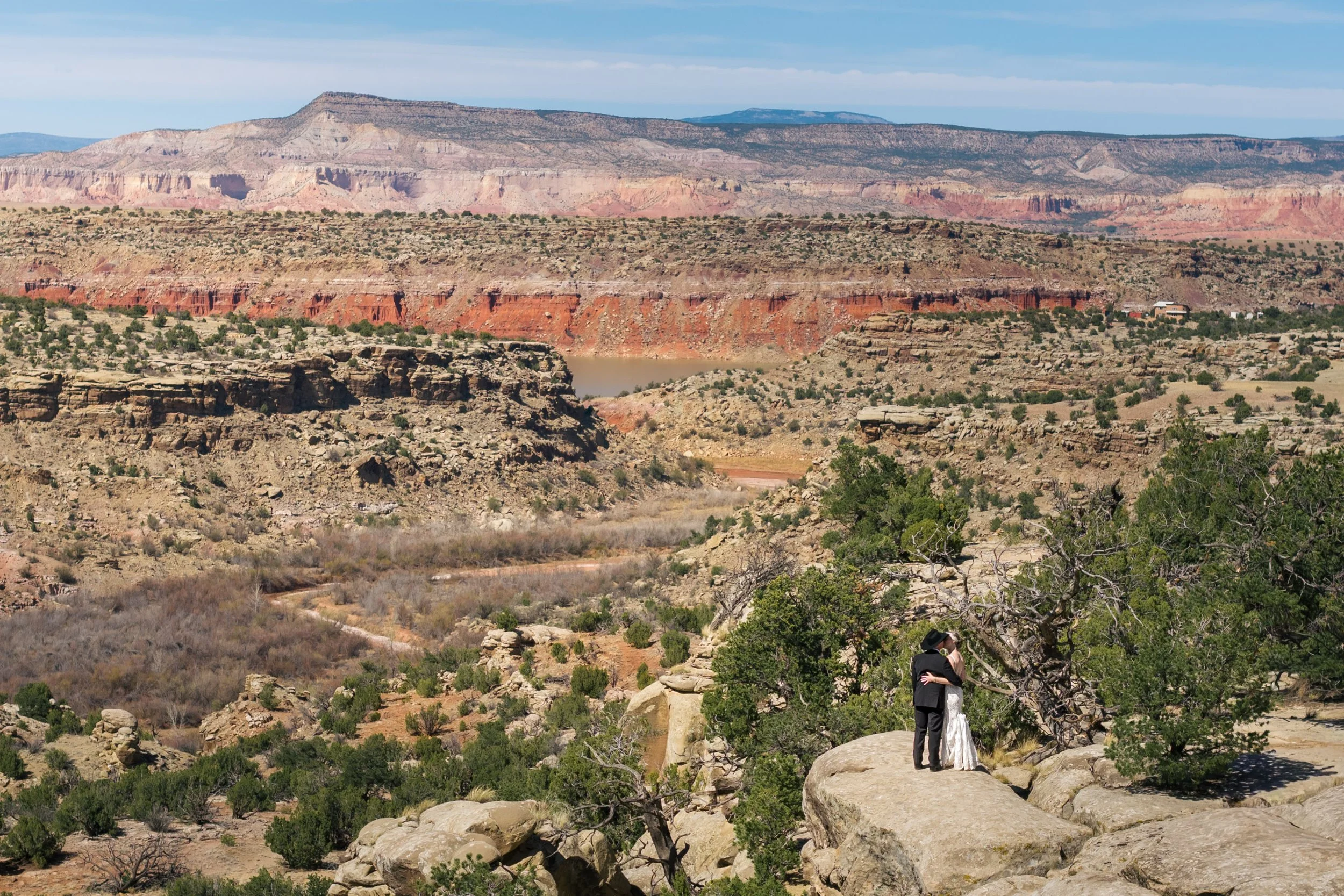 A couple dressed in wedding attire stands embracing on a rock in a desert canyon landscape with layered colorful cliffs and sparse vegetation.