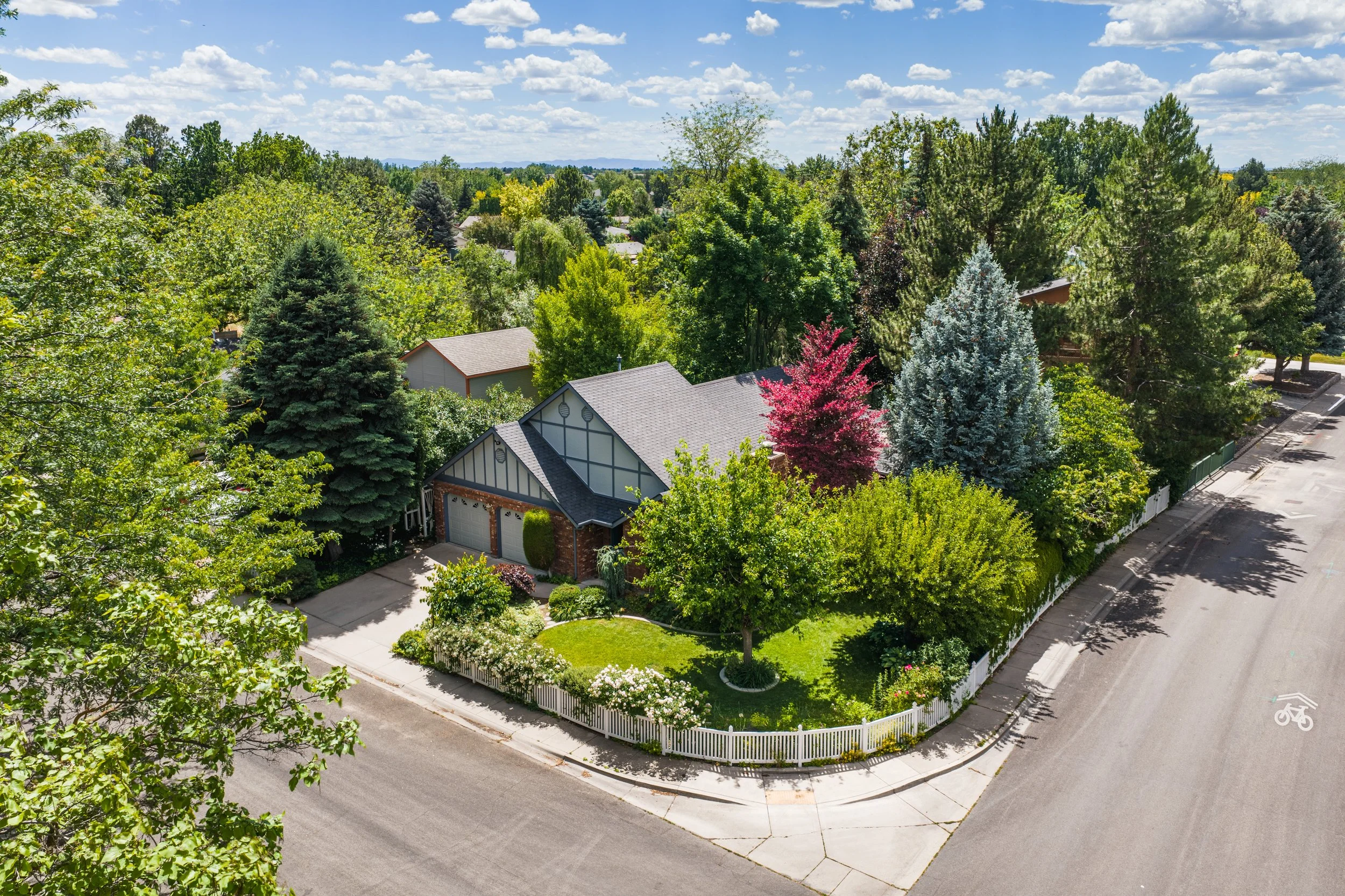 An aerial view of a suburban neighborhood with houses surrounded by lush green trees, a curved street, and a bright blue sky with scattered clouds.