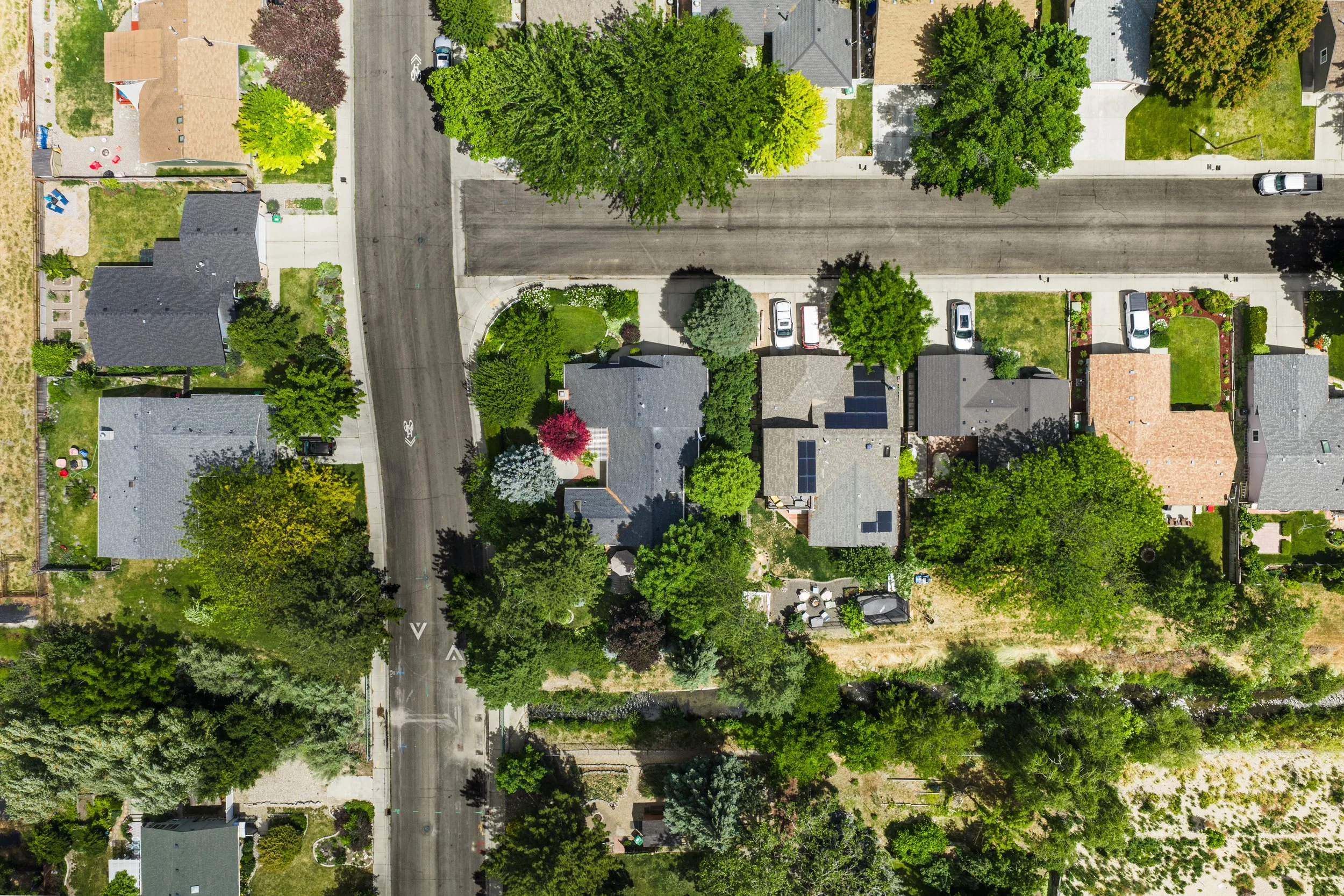 An aerial view of a suburban neighborhood with houses, trees, and parked cars along a street.