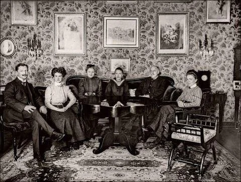 Six people sitting around a table in a vintage living room with framed artwork on wallpapered walls.