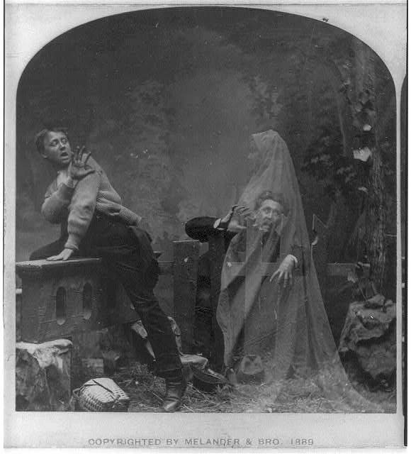 Historical black and white photograph showing three people in front of a painted backdrop with trees. One person is sitting on a platform, gesturing with a hand. Another person is kneeling, wearing a veil, and appears to be in costume. The third person is standing, probably in disguise or costume, and partly obscured. The photograph is labeled "COPYRIGHTED BY MELANDER & BRO. 1889".