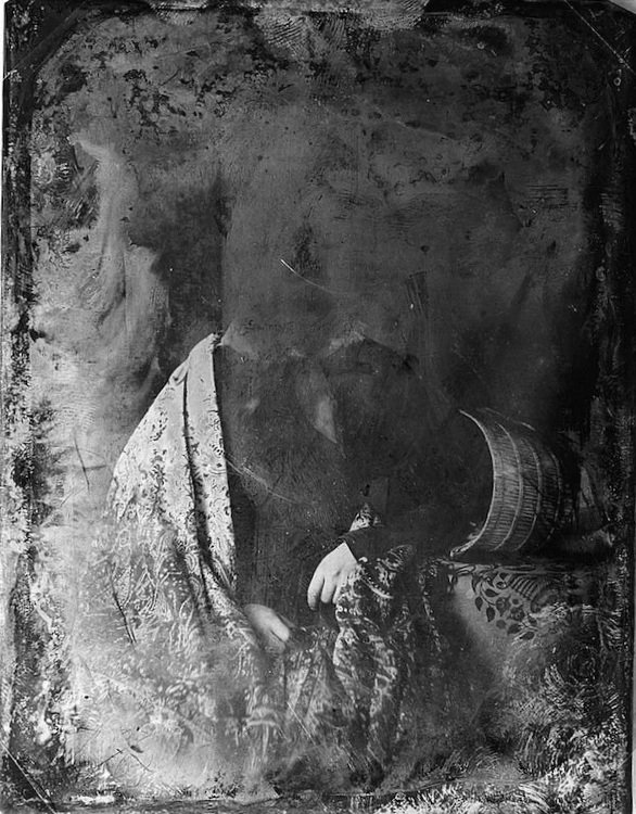 A black-and-white, damaged, or deteriorated photograph of a woman holding a book, with her face and some parts of the image obscured or faded.