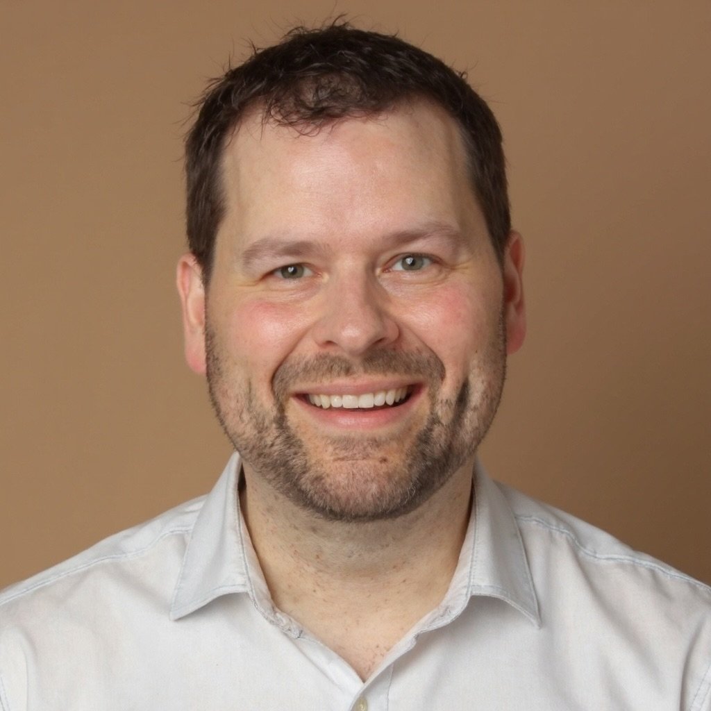 A smiling man with short brown hair and a beard, wearing a light-colored shirt, in front of a plain tan background.