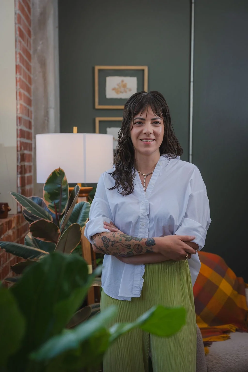 Image of a smiling latinx therapist from Wichita, Kansas standing near a large window near indoor plants. The therapist has tattoos and a septum piercing and is wearing an oversized blouse and funky pants.
