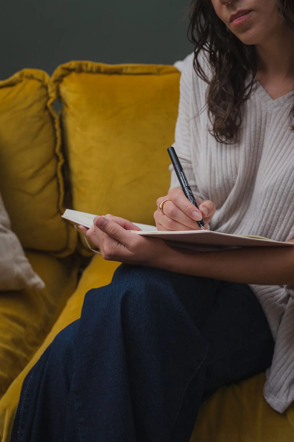 An image of an latinx Wichita, Kansas therapist writing in a notebook. The therapist is seated on a funky couch and has tattoos and a septum piercing, and is smiling warmly and welcomingly as she listens.