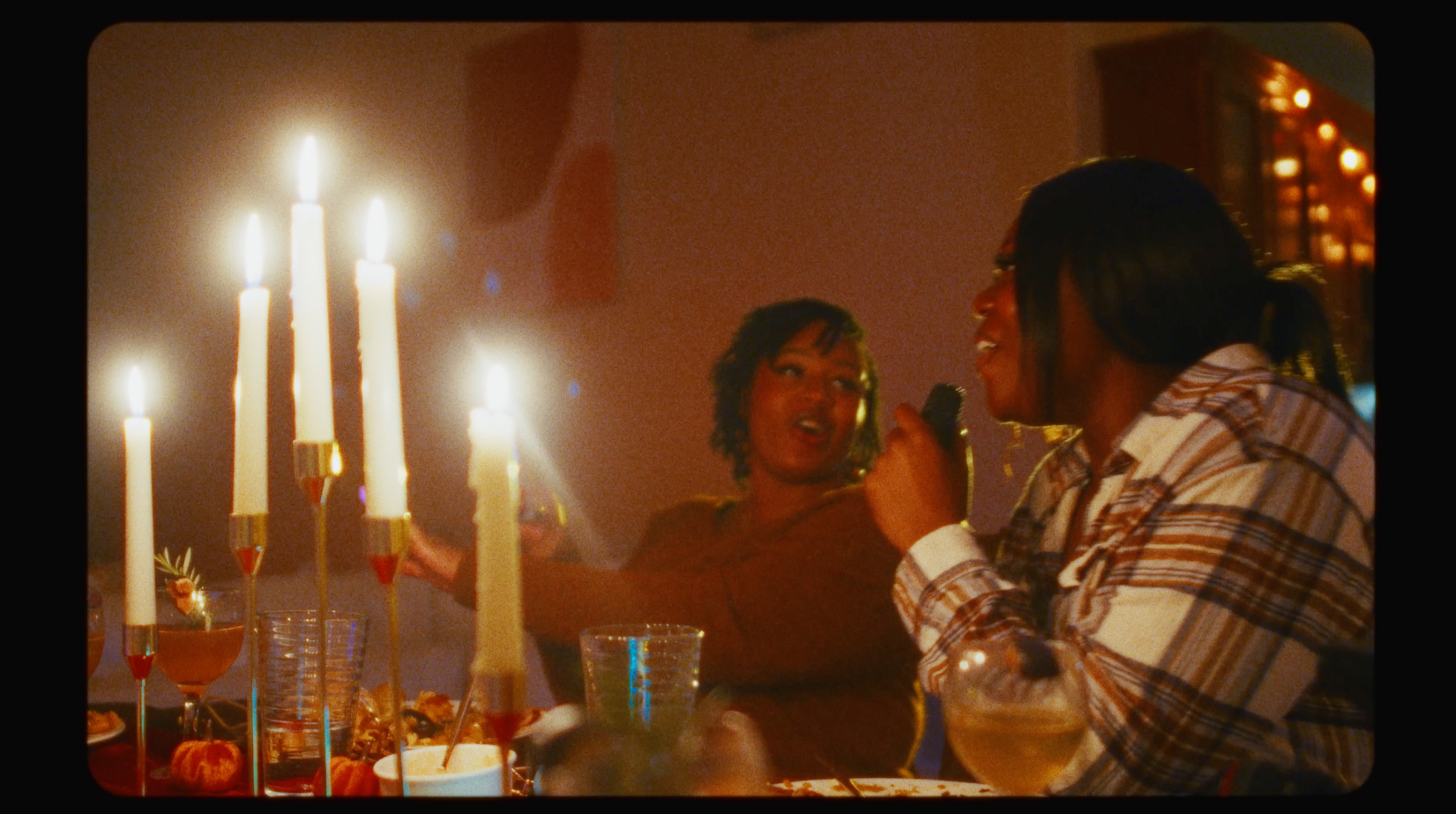 Two women are sitting at a dinner table with lit candles, enjoying conversation and drinks, in a dimly lit setting.