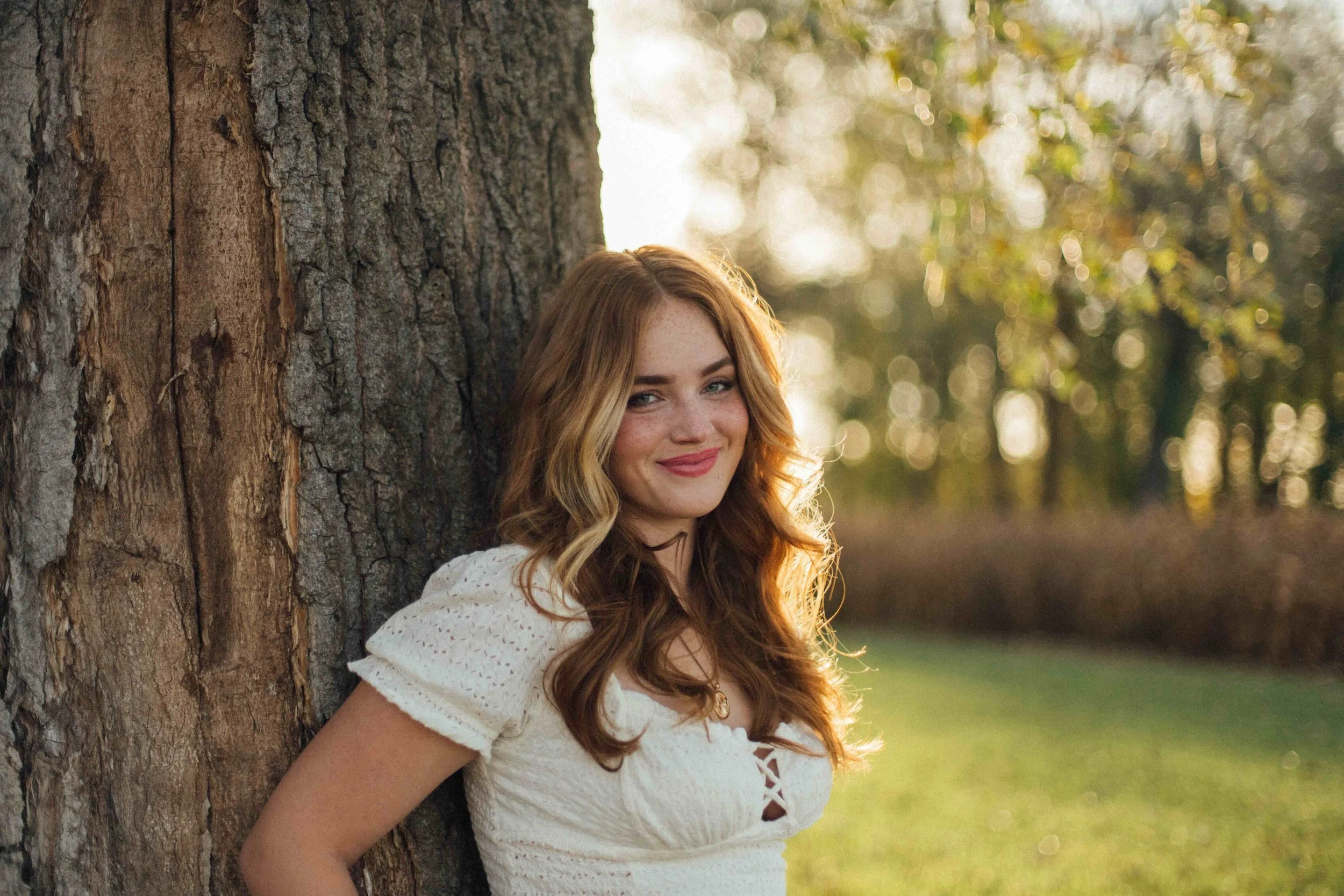 A young woman with long wavy red hair leaning against a tree trunk outdoors during golden hour, smiling, wearing a white eyelet dress, with greenery and sunlight in the background.