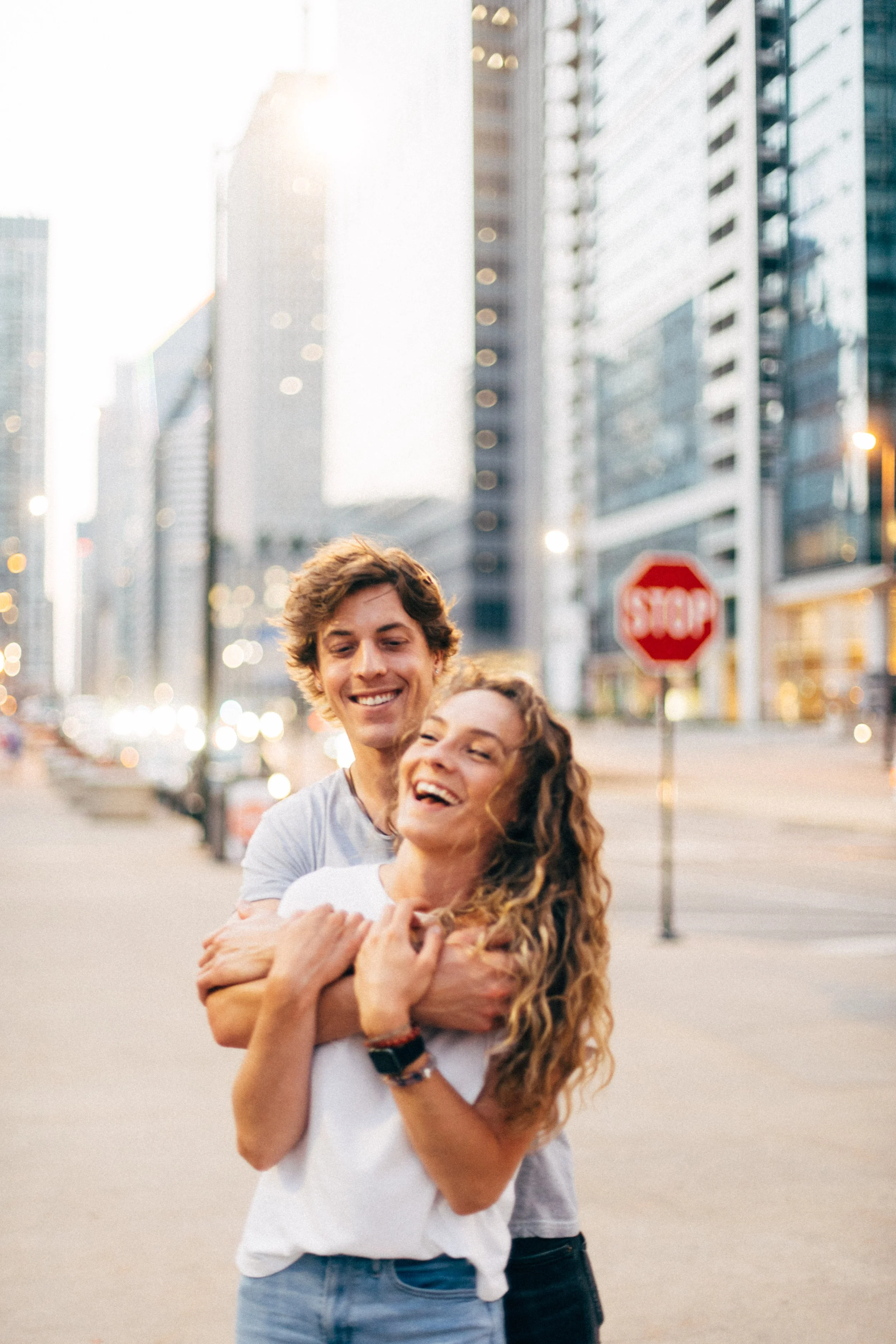 Two smiling people, a young man and woman, hugging each other on a city street during sunset. The background features tall buildings and a stop sign.