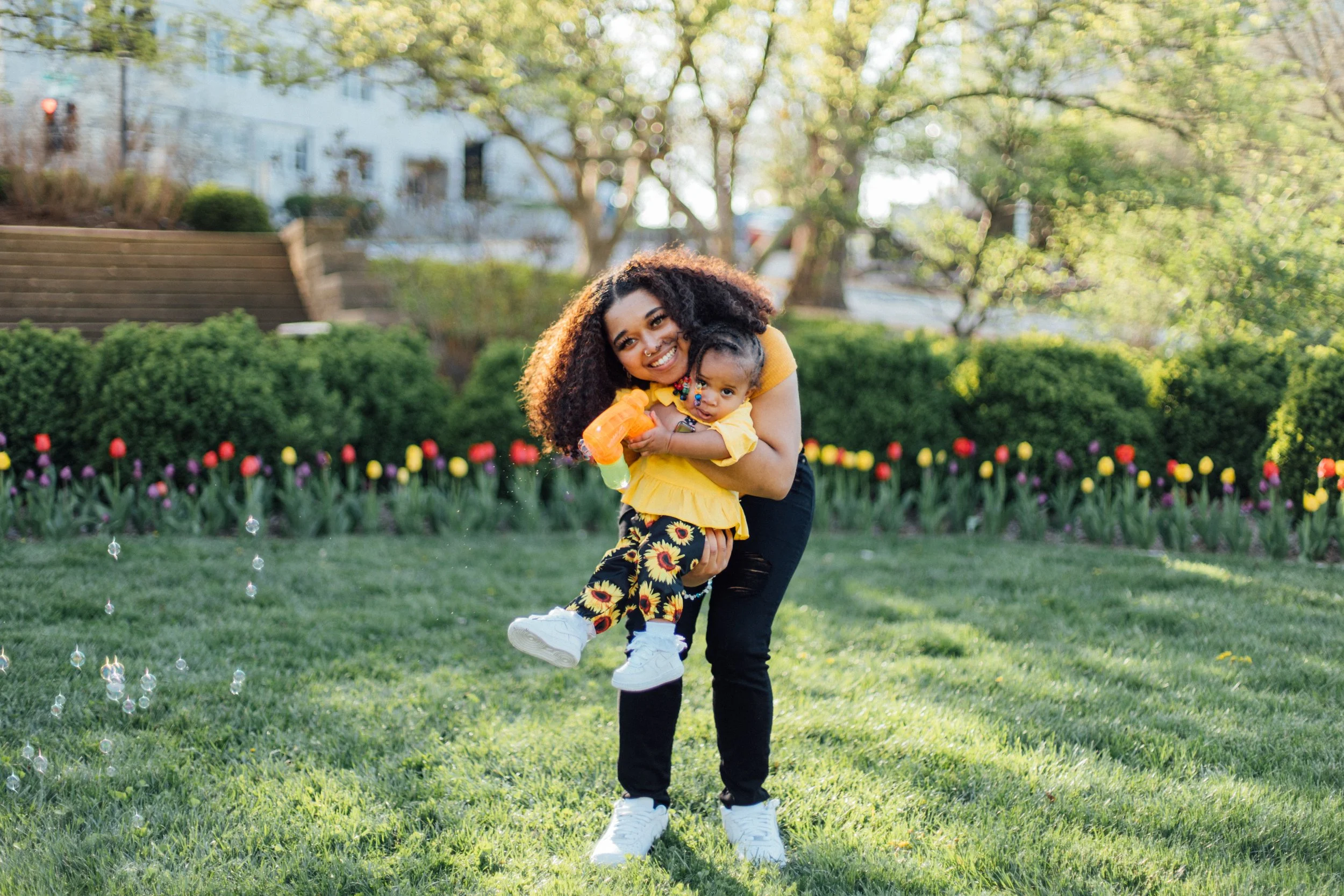 A woman with curly hair smiling and holding a young girl in a yellow outfit in a garden with blooming tulips, trees, and sunlight.