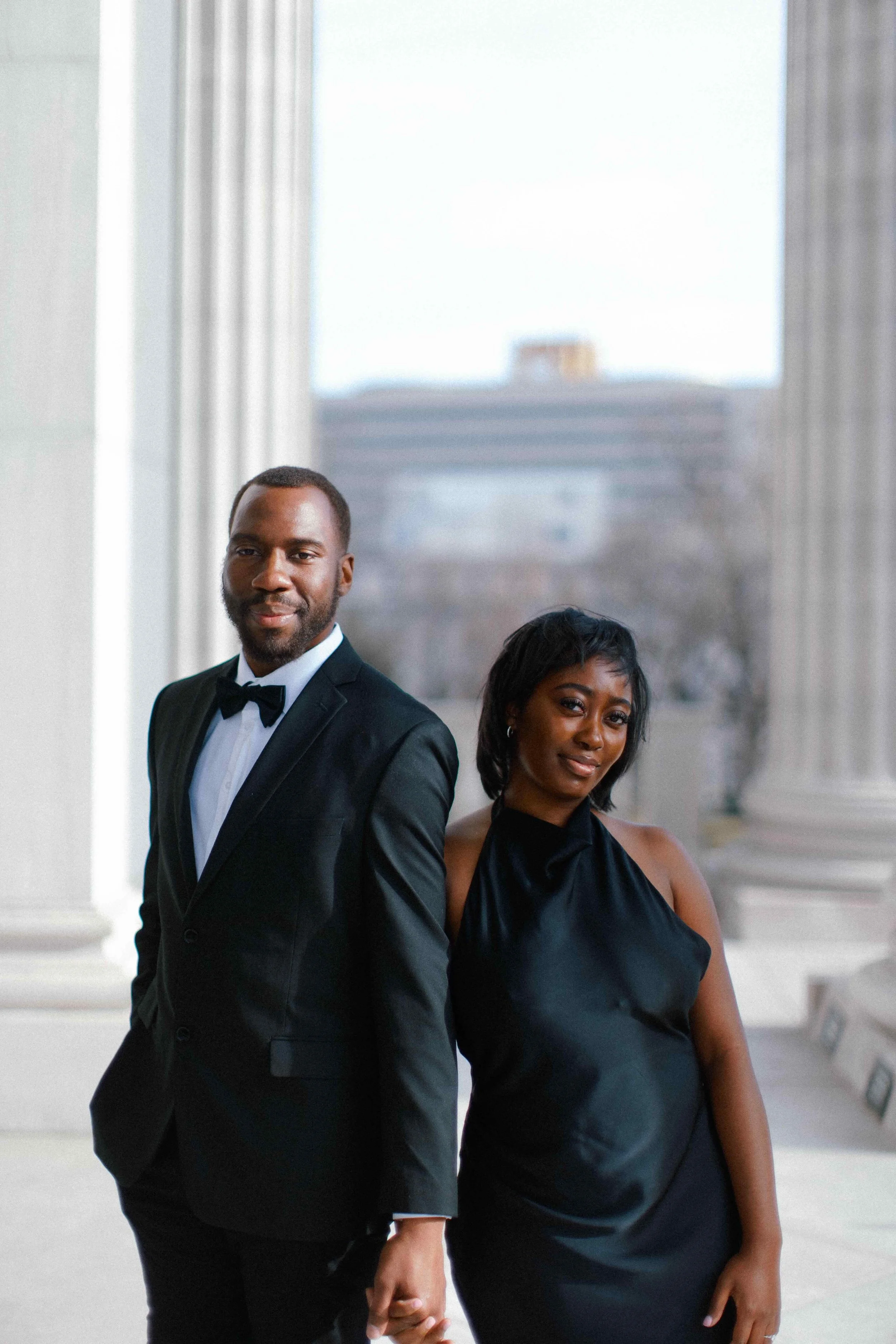 A man in a tuxedo and a woman in a black dress holding hands and standing in an elegant room.