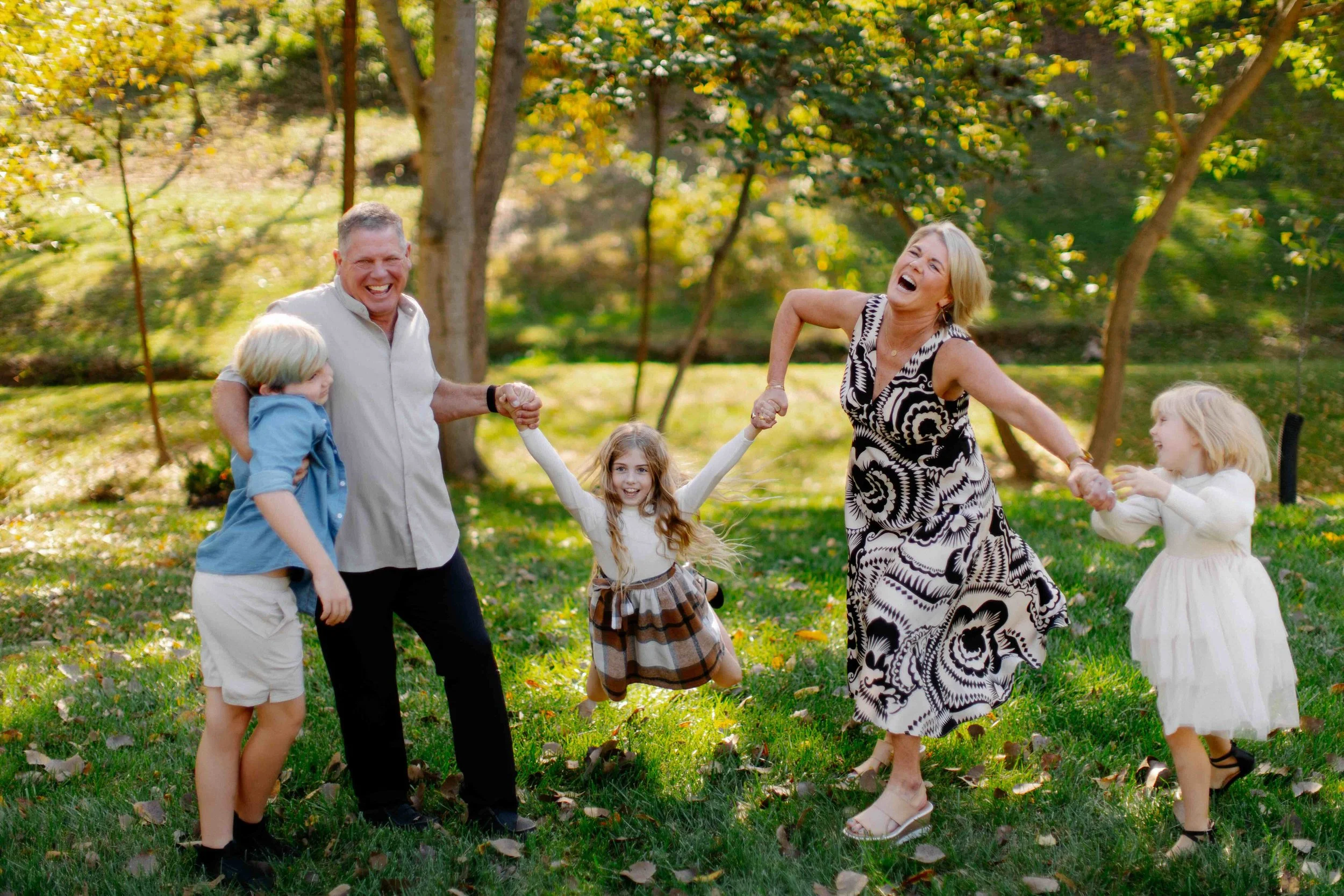 A group of five people, three children and two adults, playing in a park with green grass and trees with autumn leaves, holding hands and laughing.