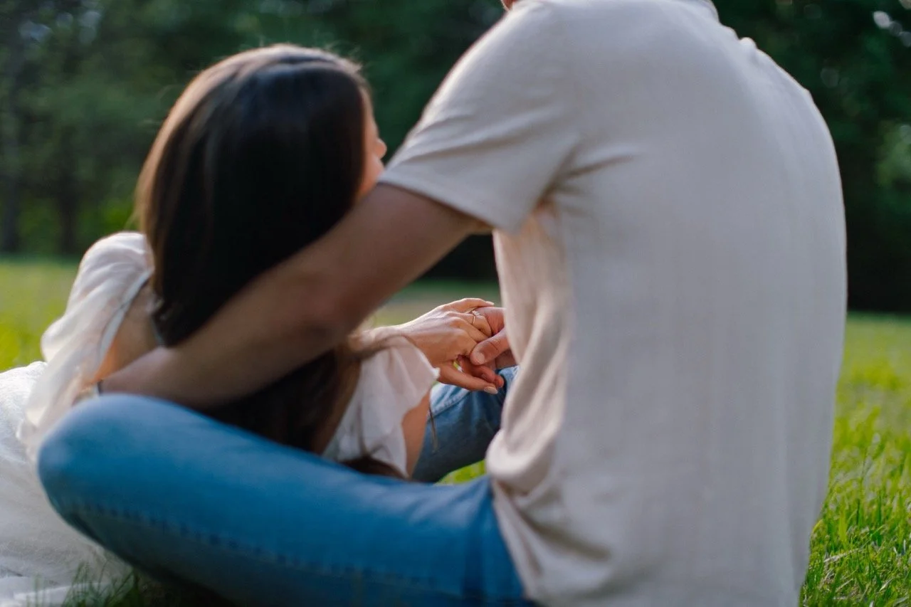 A woman and a man sitting on grass in a park, holding hands and enjoying an outdoor moment together.