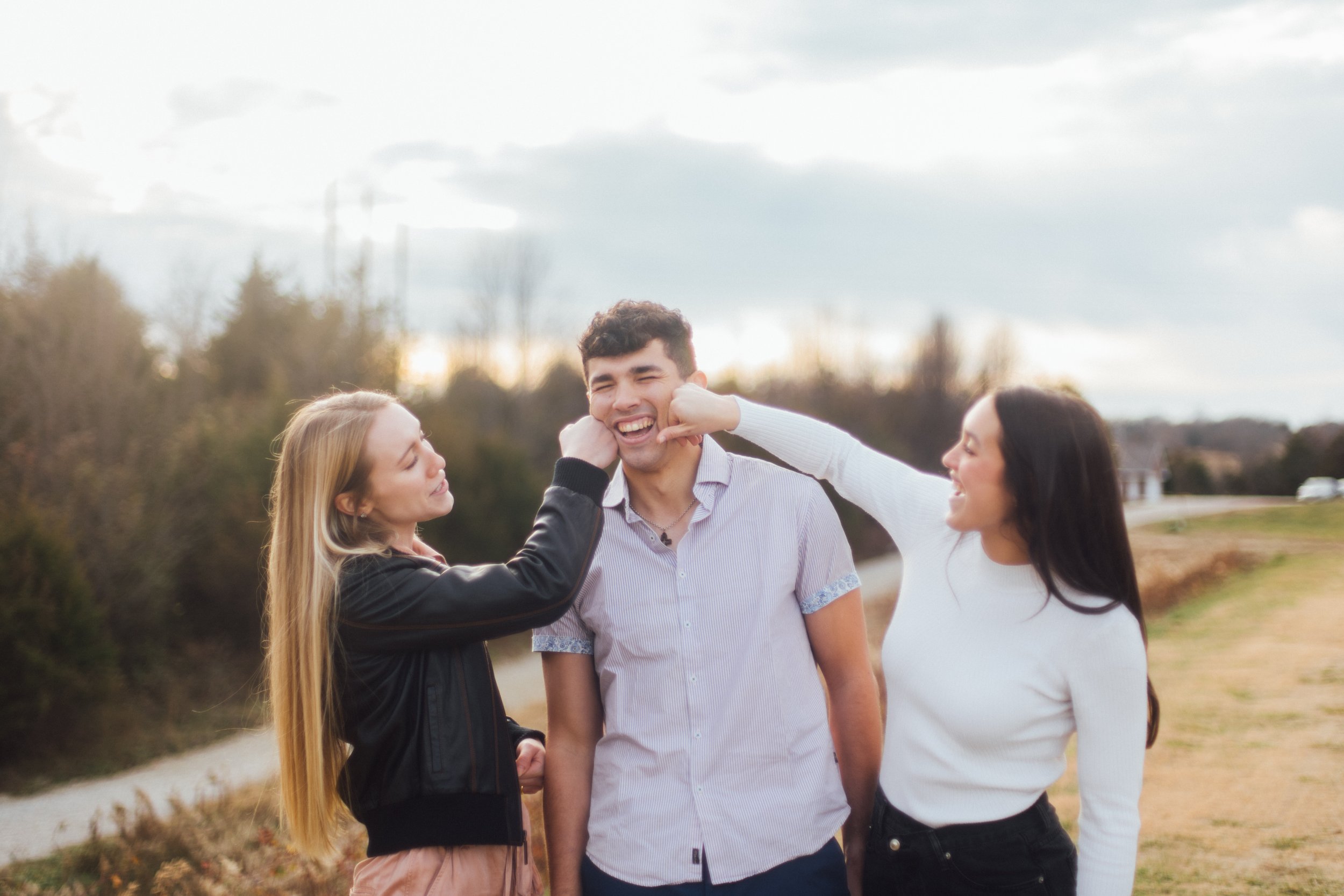 Three young women and a young man smiling outdoors on a cloudy day, with trees and a path in the background.