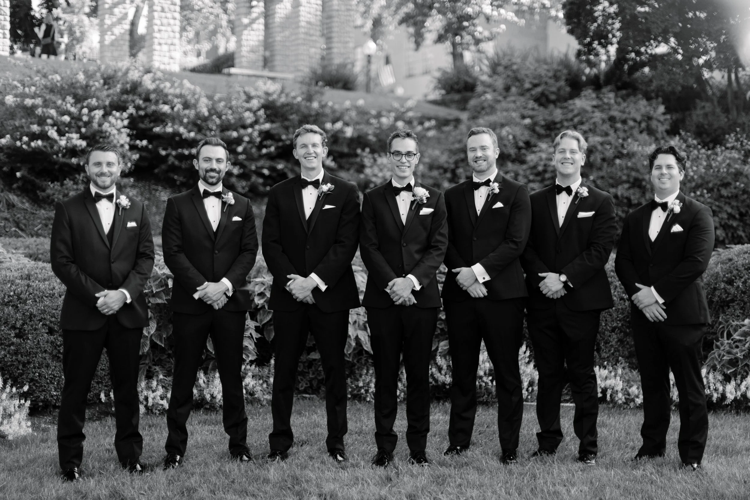 Black and white photo of seven men dressed in tuxedos with bow ties, standing outdoors in a line on grass, smiling at the camera, with trees and a building in the background.