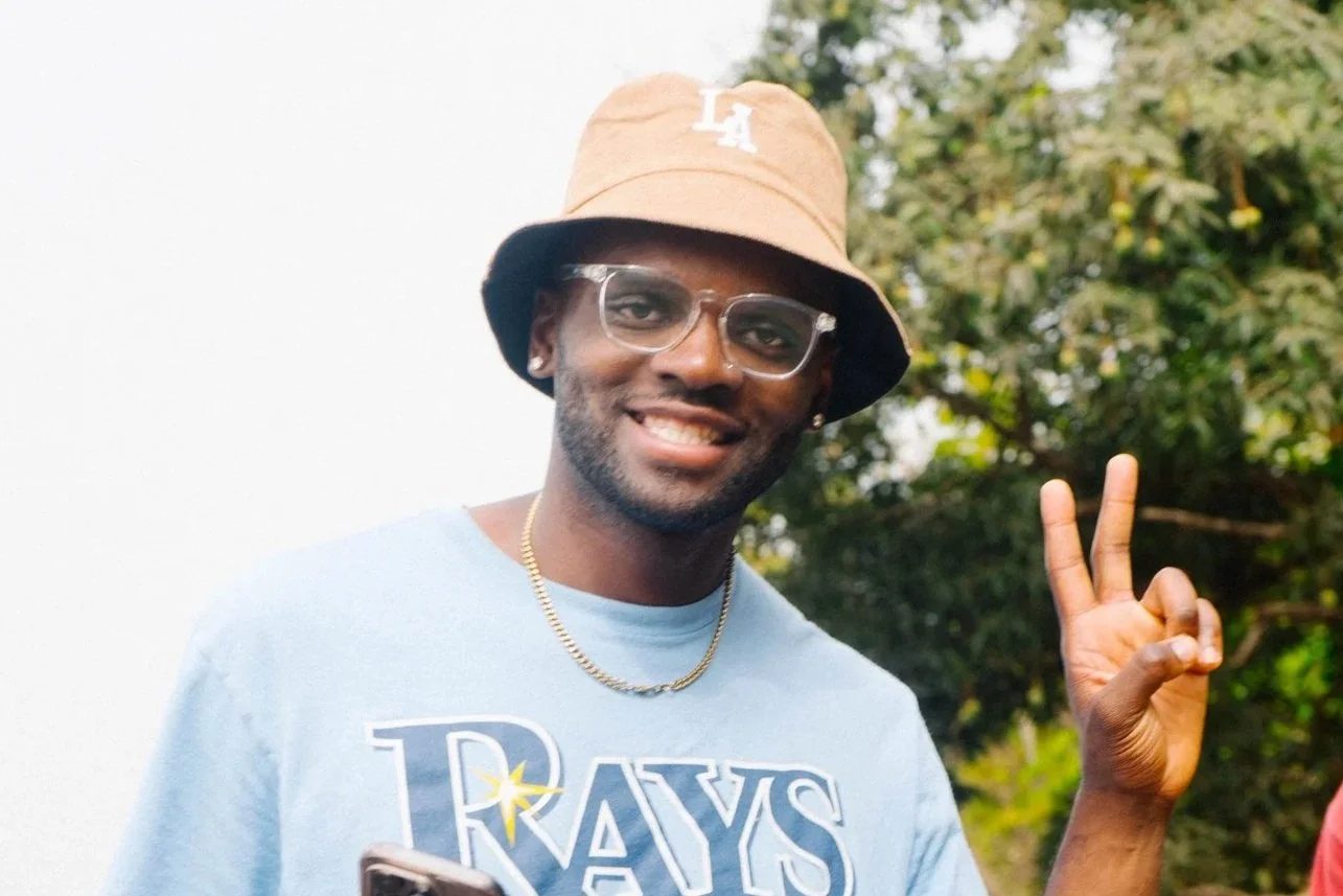 A smiling man wearing clear glasses, a tan bucket hat with 'LA' embroidery, a light blue T-shirt with 'RAYS' printed on it, and a gold chain necklace, making a peace sign with his right hand outdoors.