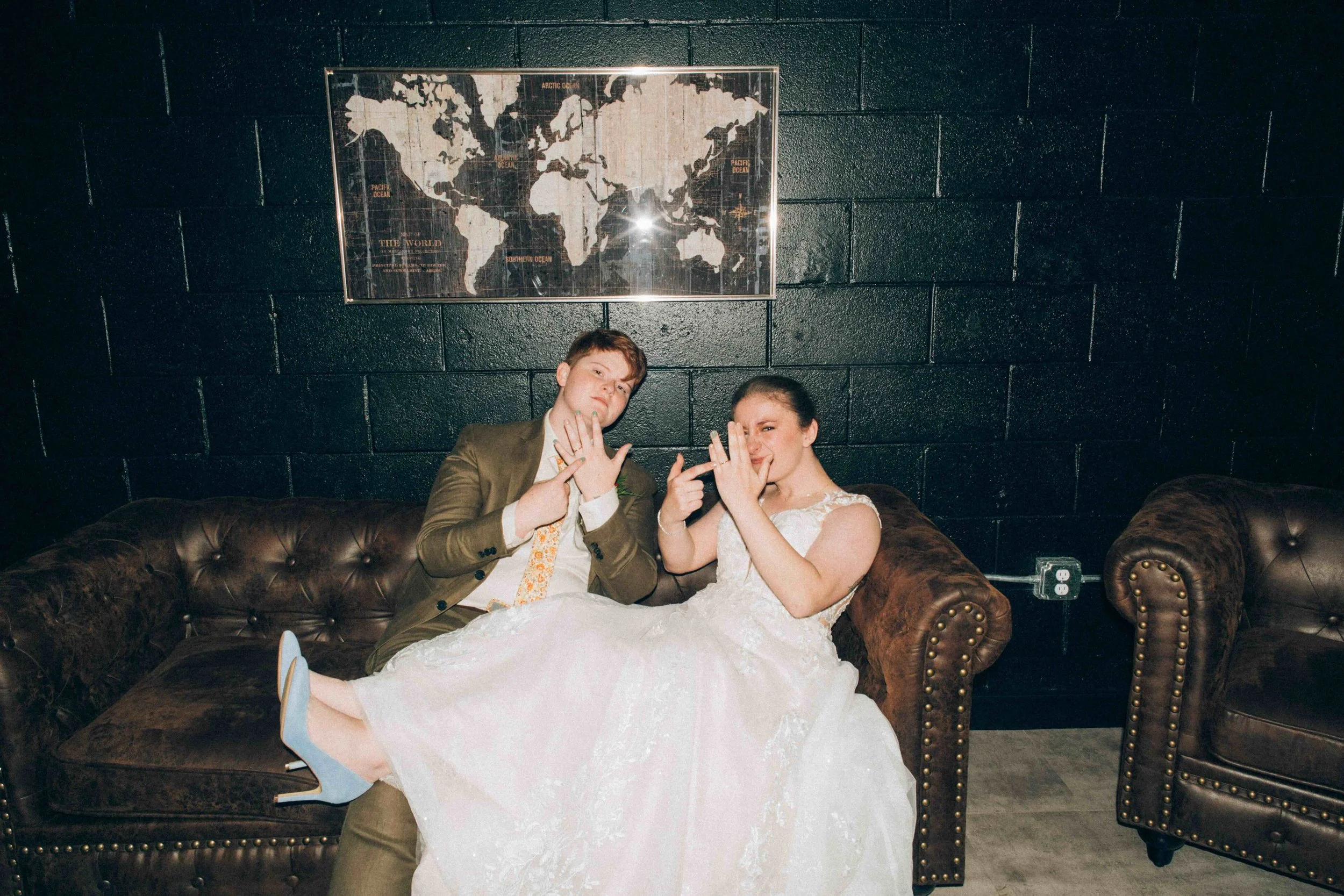 A man in a brown suit and a woman in a wedding dress are sitting on a dark brown leather couch against a dark wall with a world map. They are making playful hand gestures and smiling.