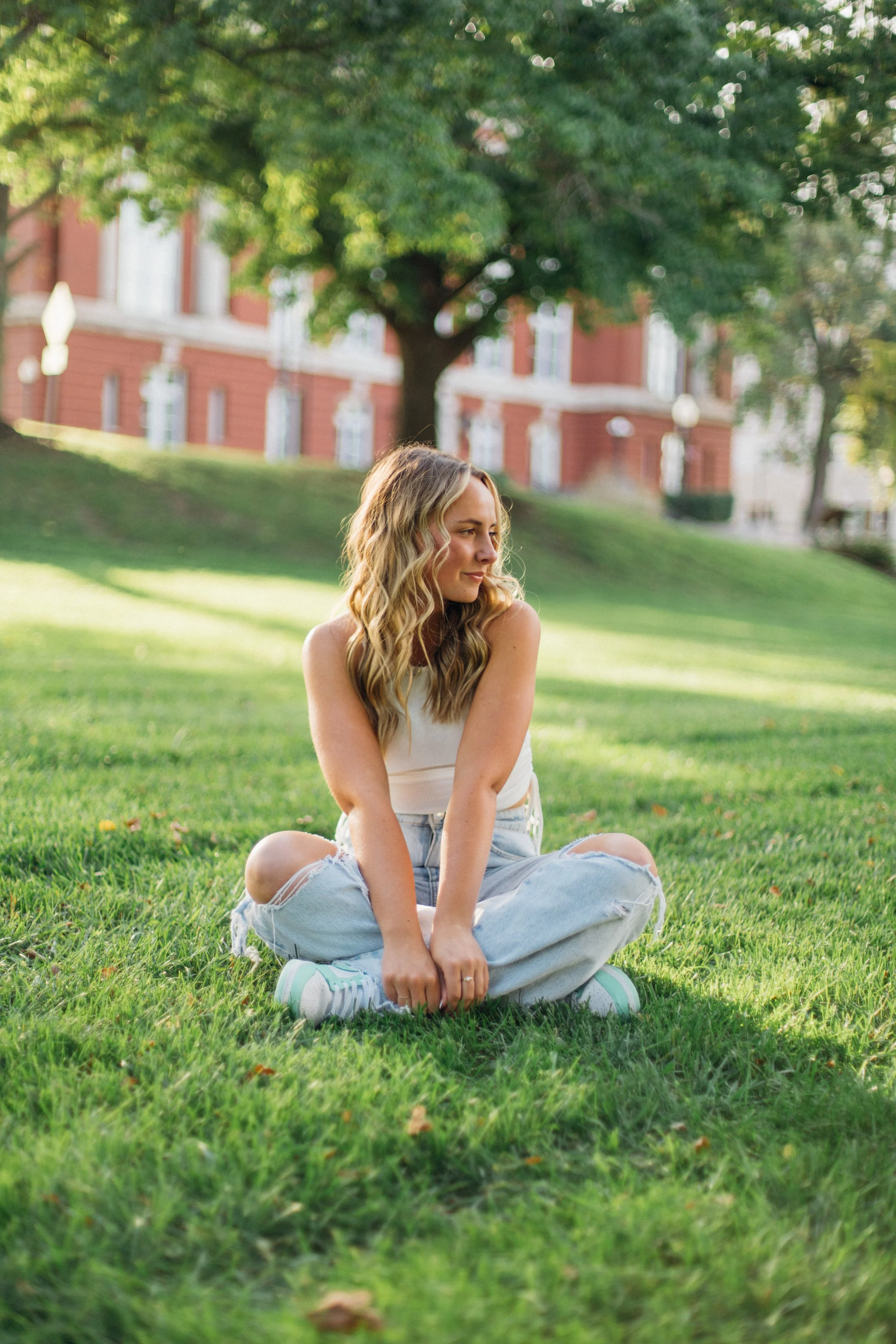 A woman with long wavy blonde hair sits cross-legged on grass in a park. She is looking to her right and smiling slightly. She wears a white tank top, ripped jeans, and sneakers. In the background, there are trees and a red brick building.