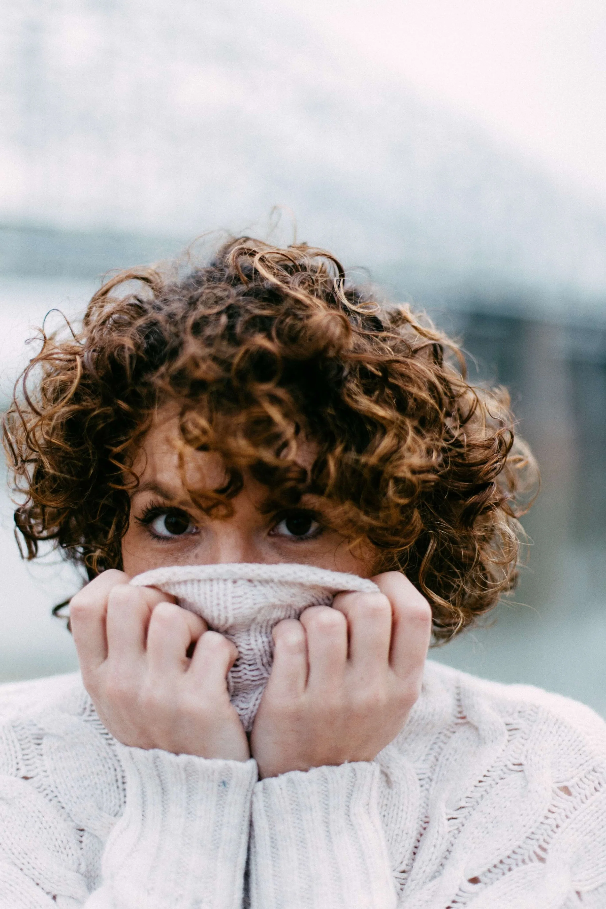 A woman with curly hair holds a beige knit sweater over her nose and mouth, peering out with wide eyes. She is wearing a cream-colored sweater.