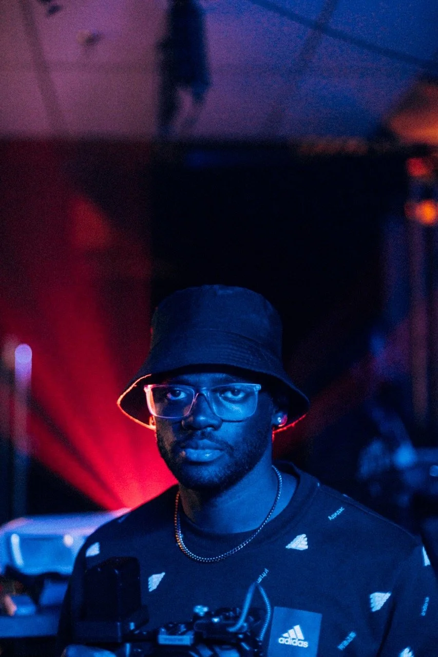 A man with glasses wearing a bucket hat and a black Adidas shirt in a dimly lit room with red and blue lighting.