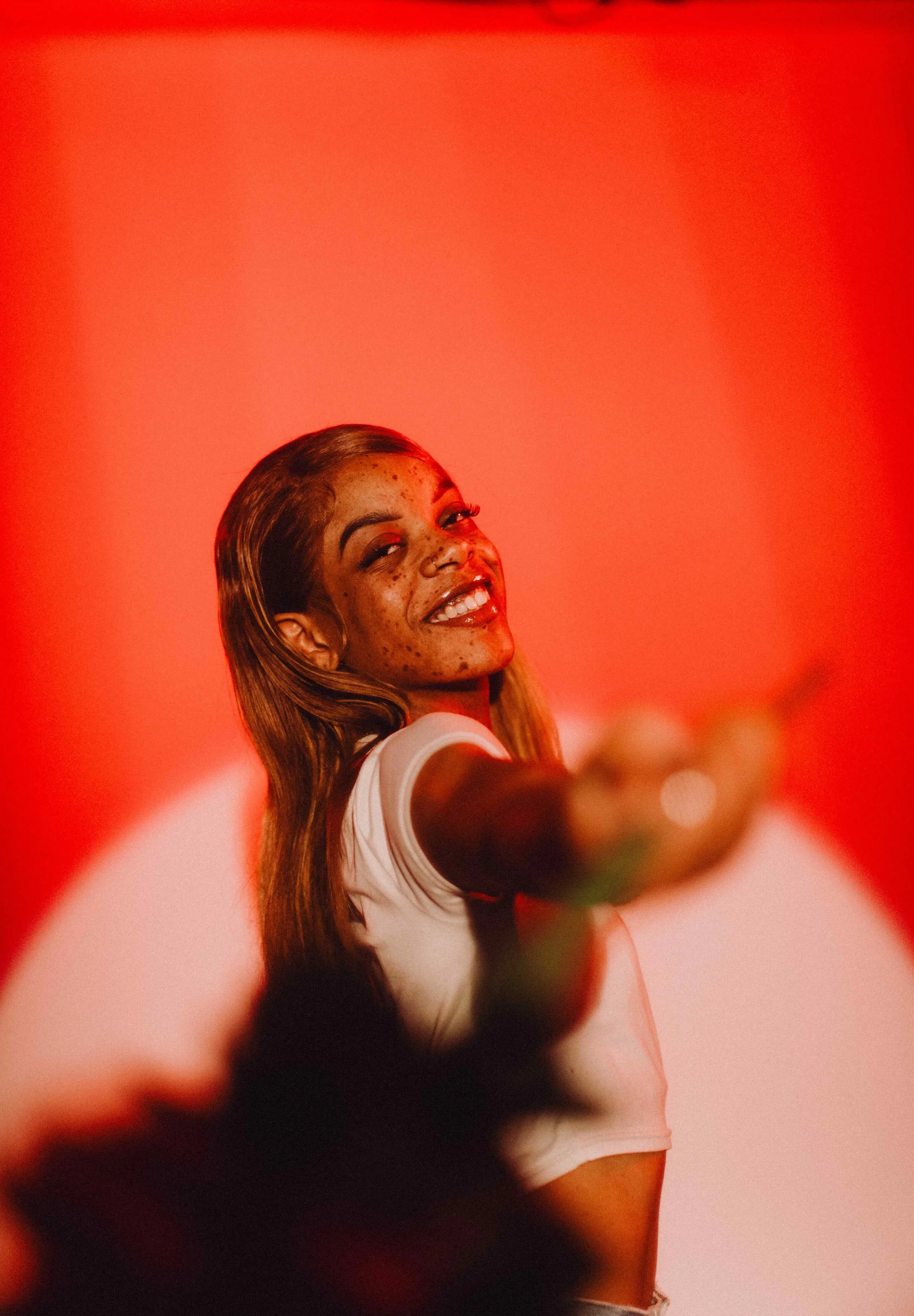 Smiling young woman with freckles and long hair, taking a selfie against a red gradient background.