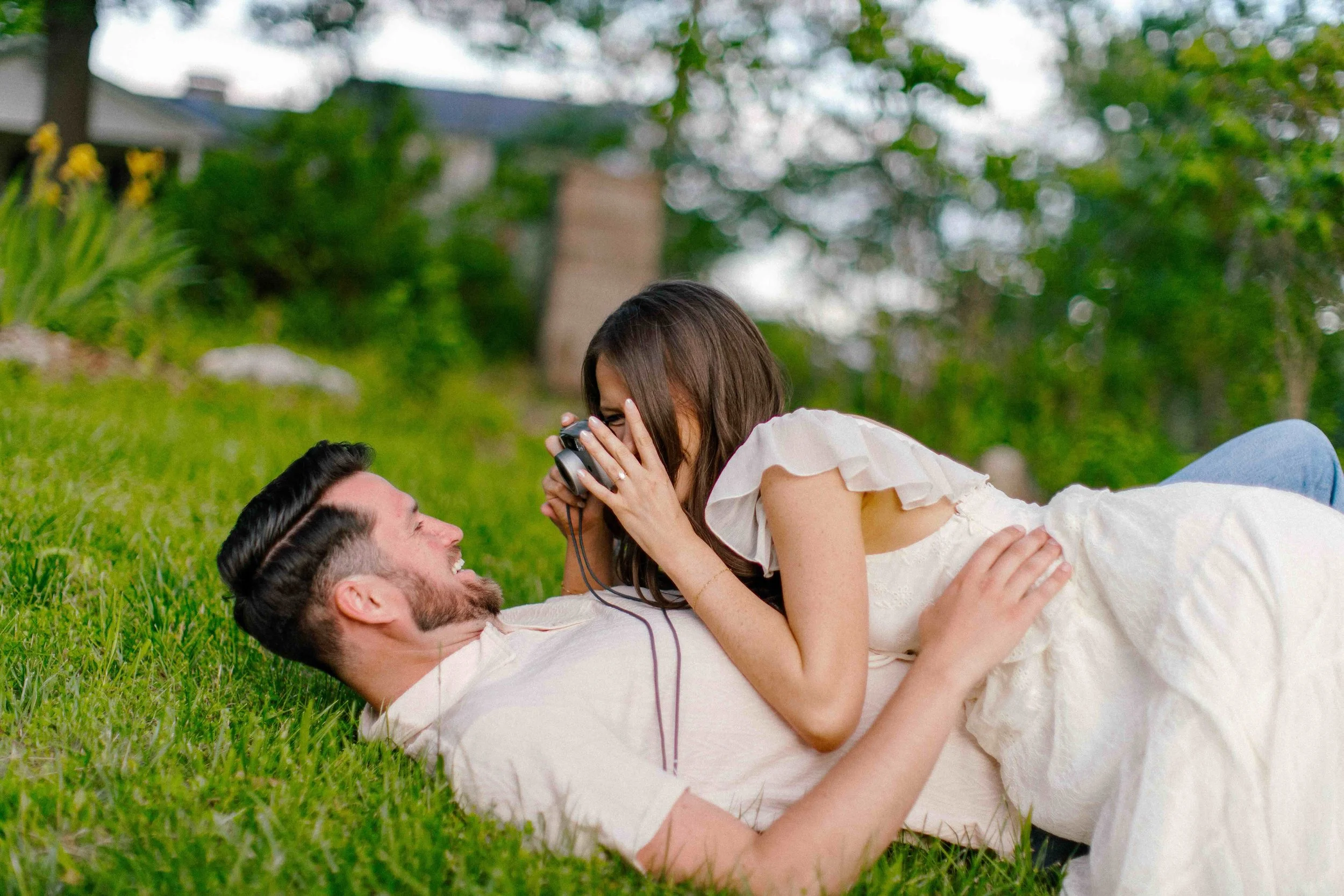 A young couple lying on grass outdoors, the woman on top of the man, both smiling and looking into each other's eyes, with the woman holding a phone to her face.