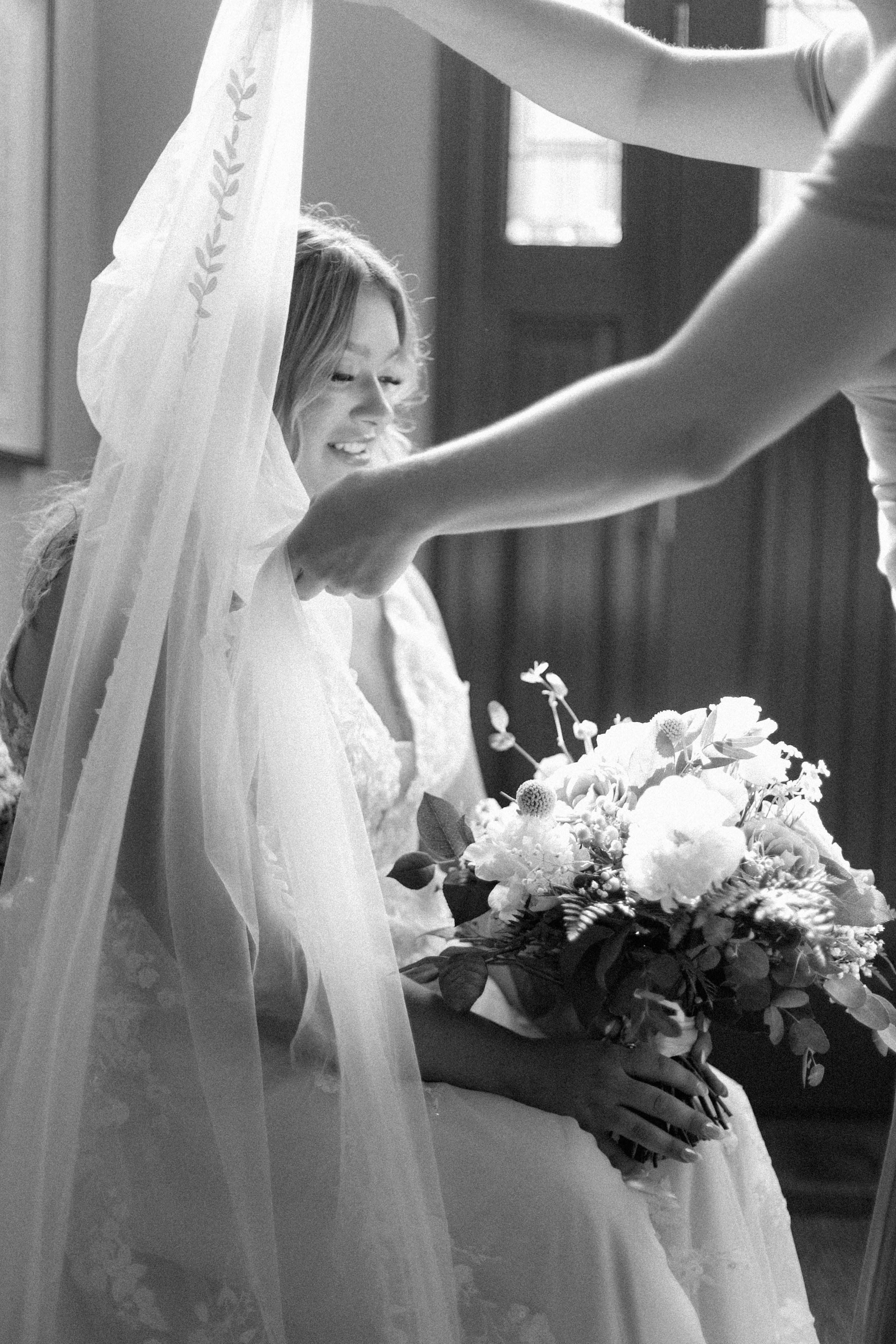A bride sitting with a big bouquet of flowers, smiling softly as a person, possibly an attendant or florist, adjusts her veil or dress.