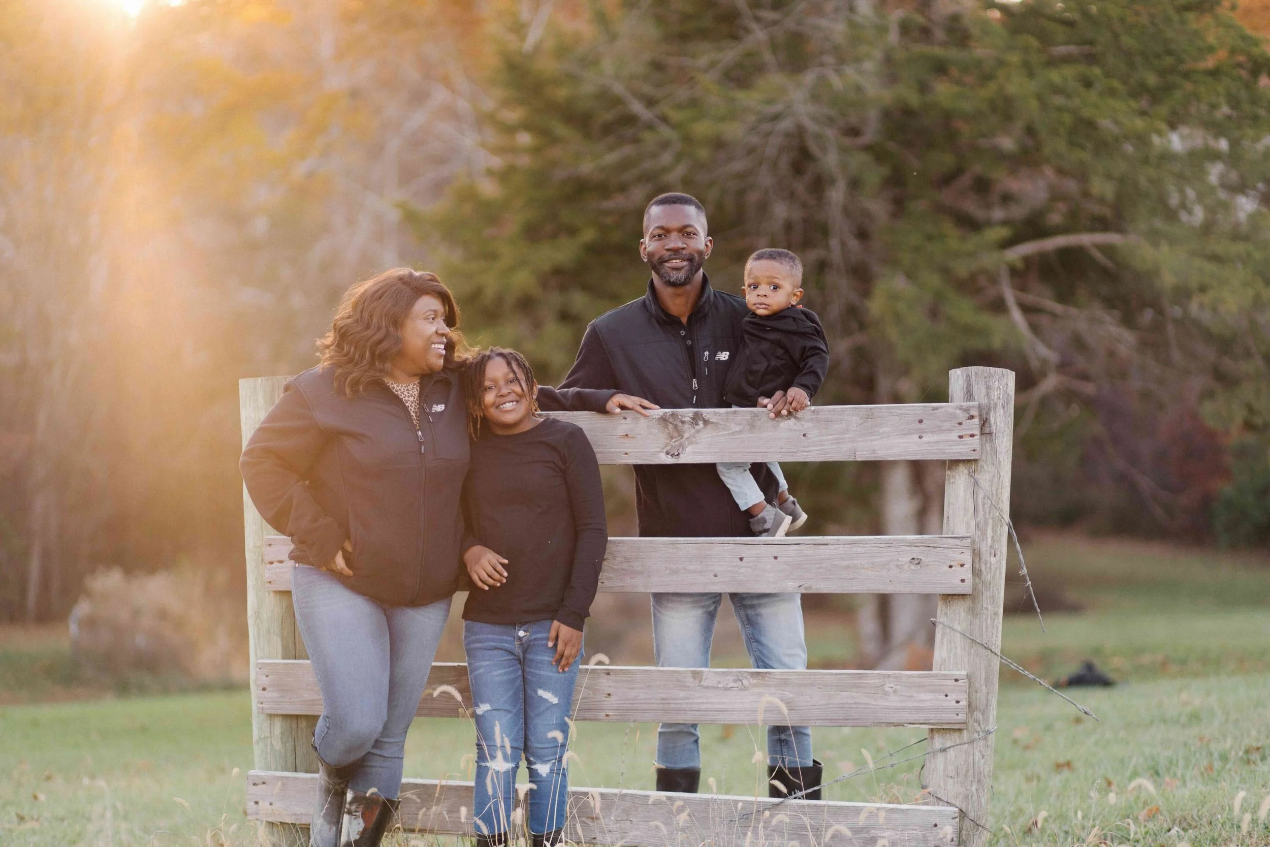 A family of four standing outdoors near a wooden fence with trees in the background during fall.