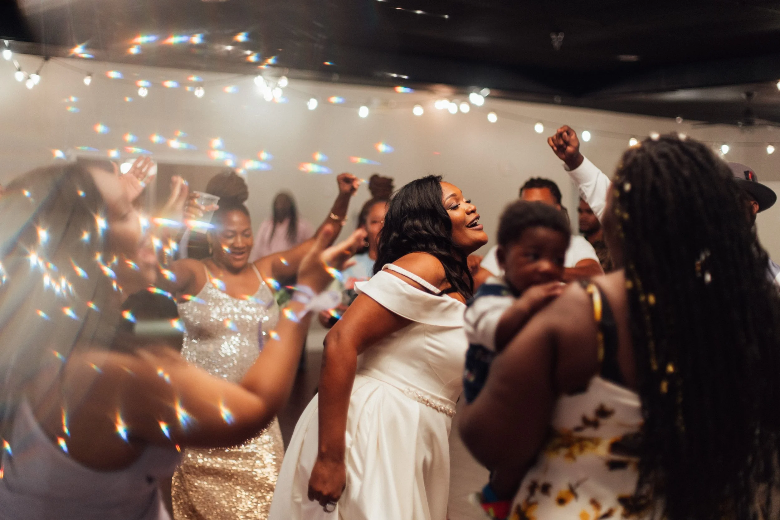 Group of people dancing and celebrating at a party, smiling and raising their hands, with festive lighting and decorations in the background.