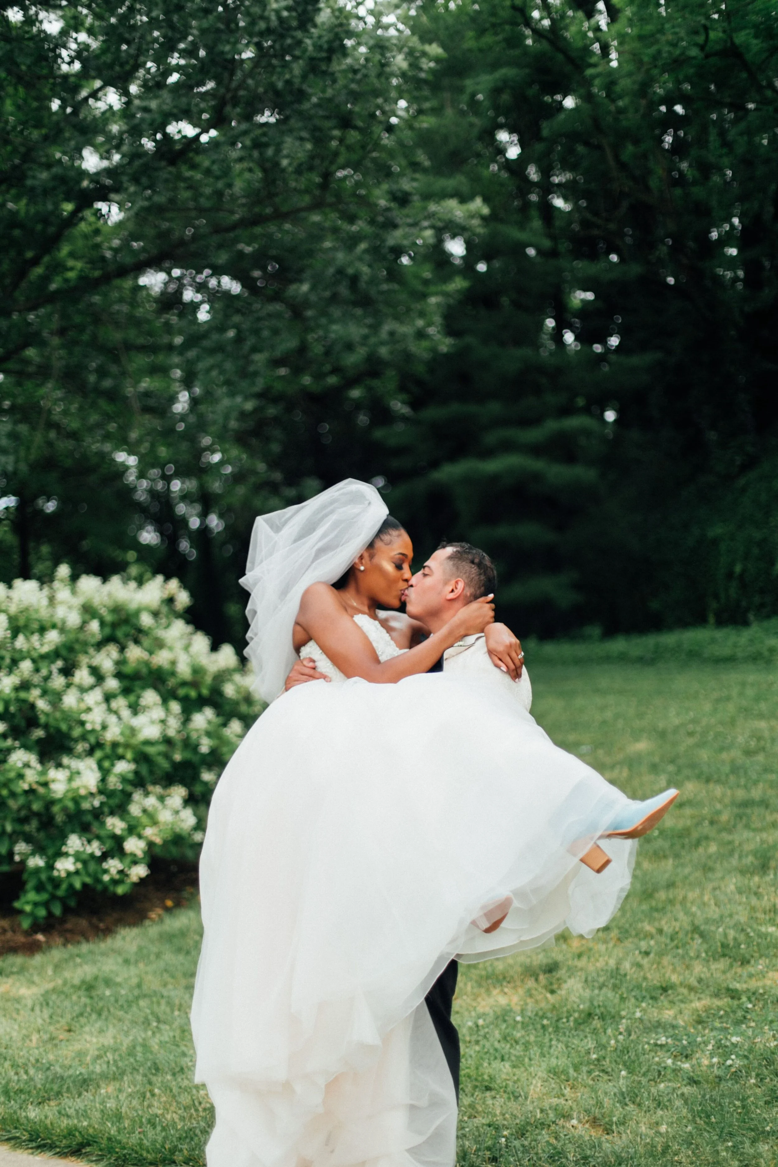 A bride and groom sharing a kiss outdoors among trees, with the bride wearing a veil and holding the groom in his arms.