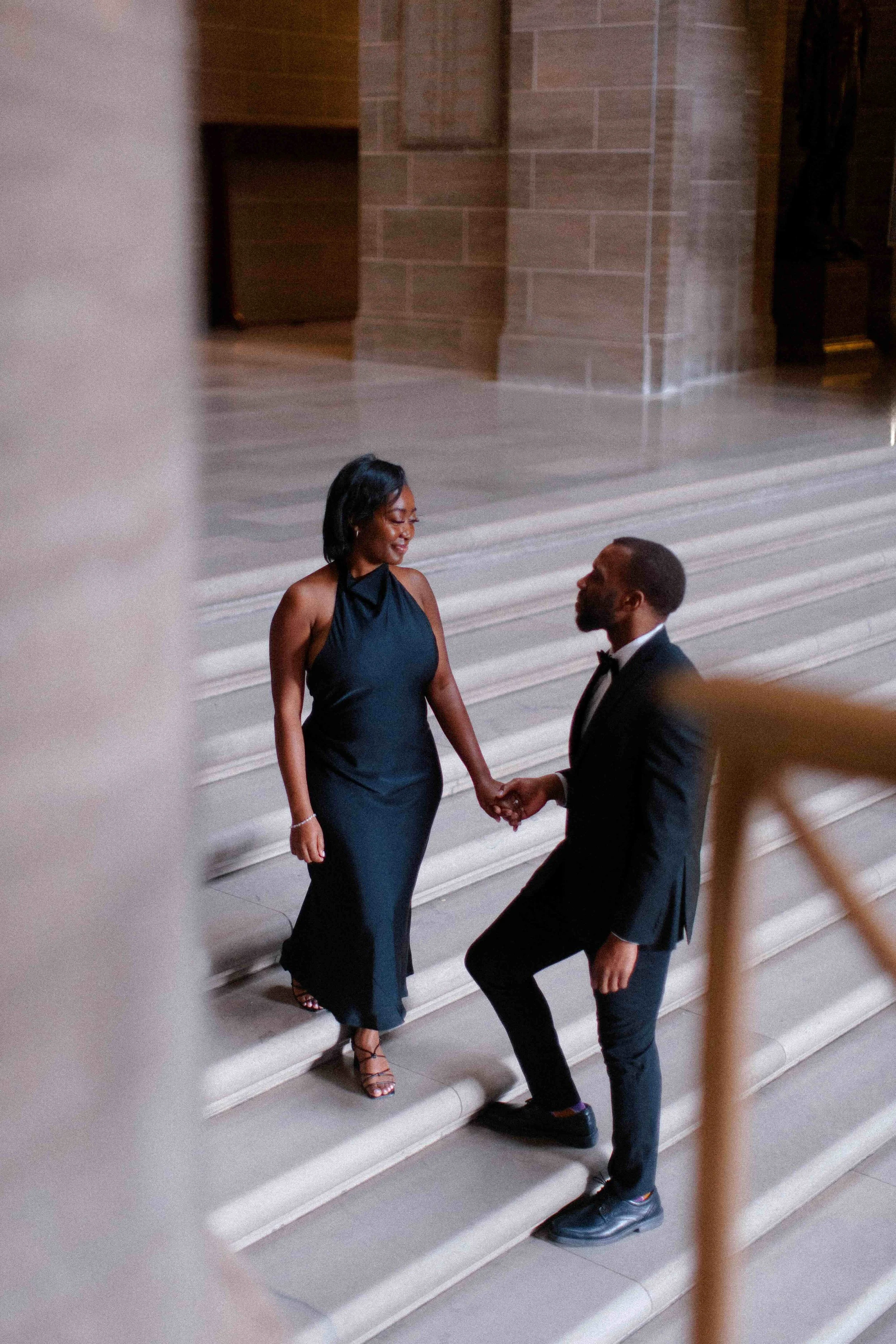 A man kneeling on a staircase holding hands with a woman standing on the stairs, both dressed in formal wear, smiling and looking at each other.