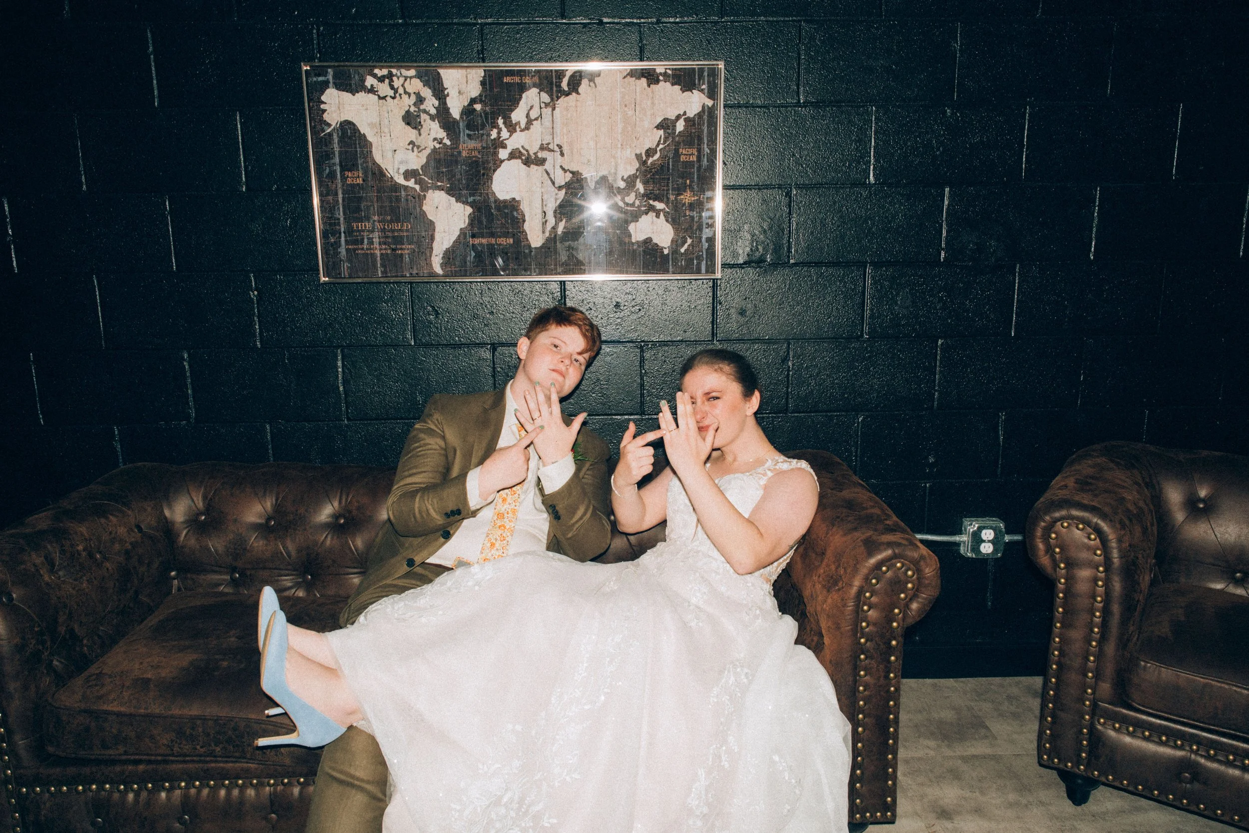 A young man in a brown suit and a young woman in a white wedding dress sit on a dark brown leather couch with button tufting, striking playful poses with their hands near their faces. Behind them is a black brick wall with a framed world map.