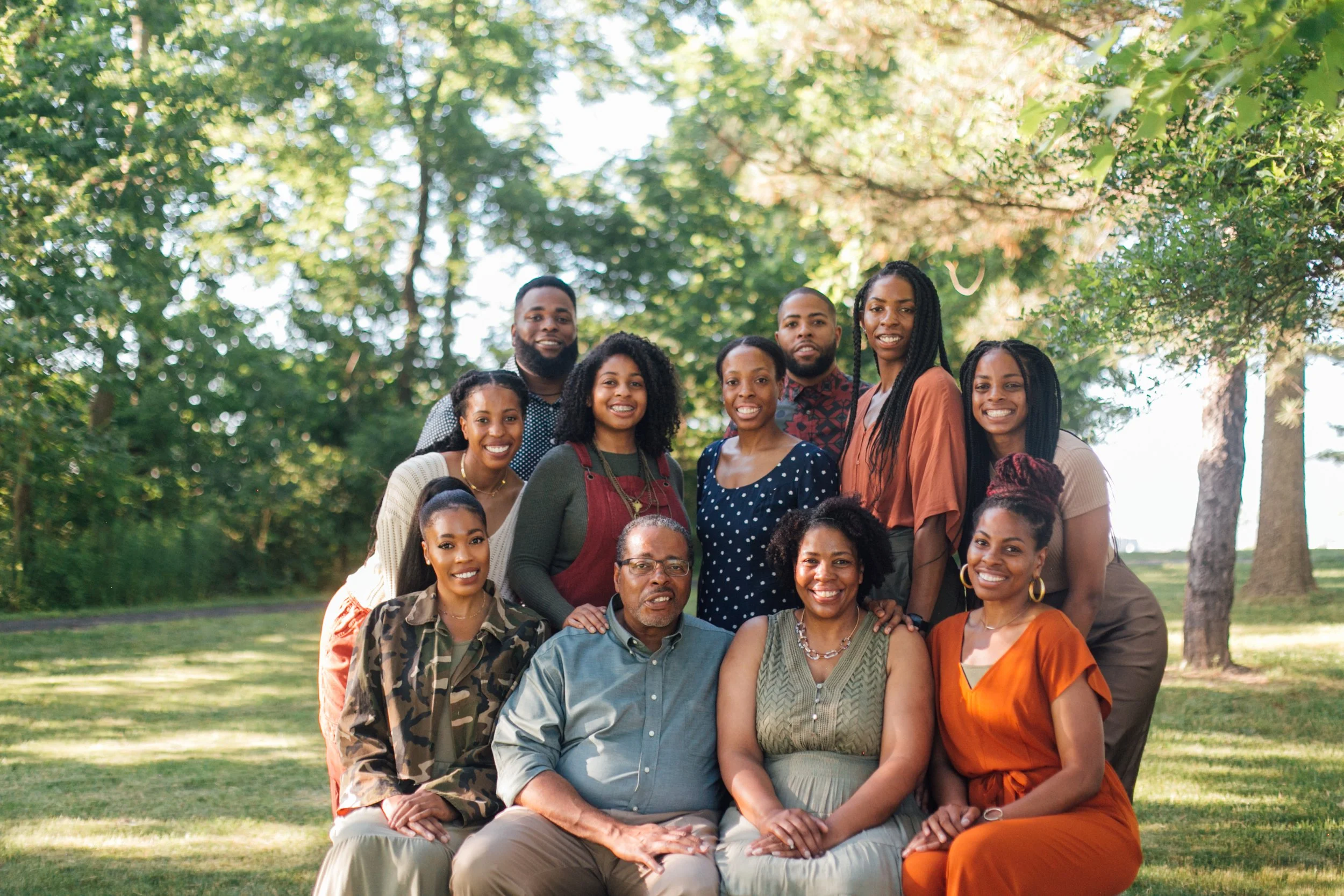 A diverse group of smiling people posing outdoors in a park with green trees in the background.