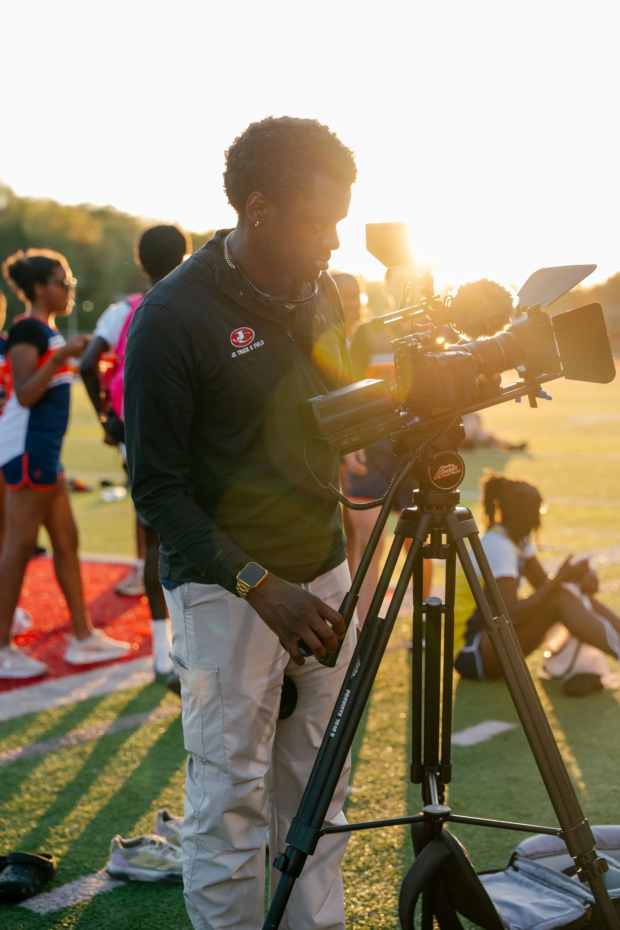 A person operating a professional video camera on a tripod at a soccer field during sunset, with teammates in the background.