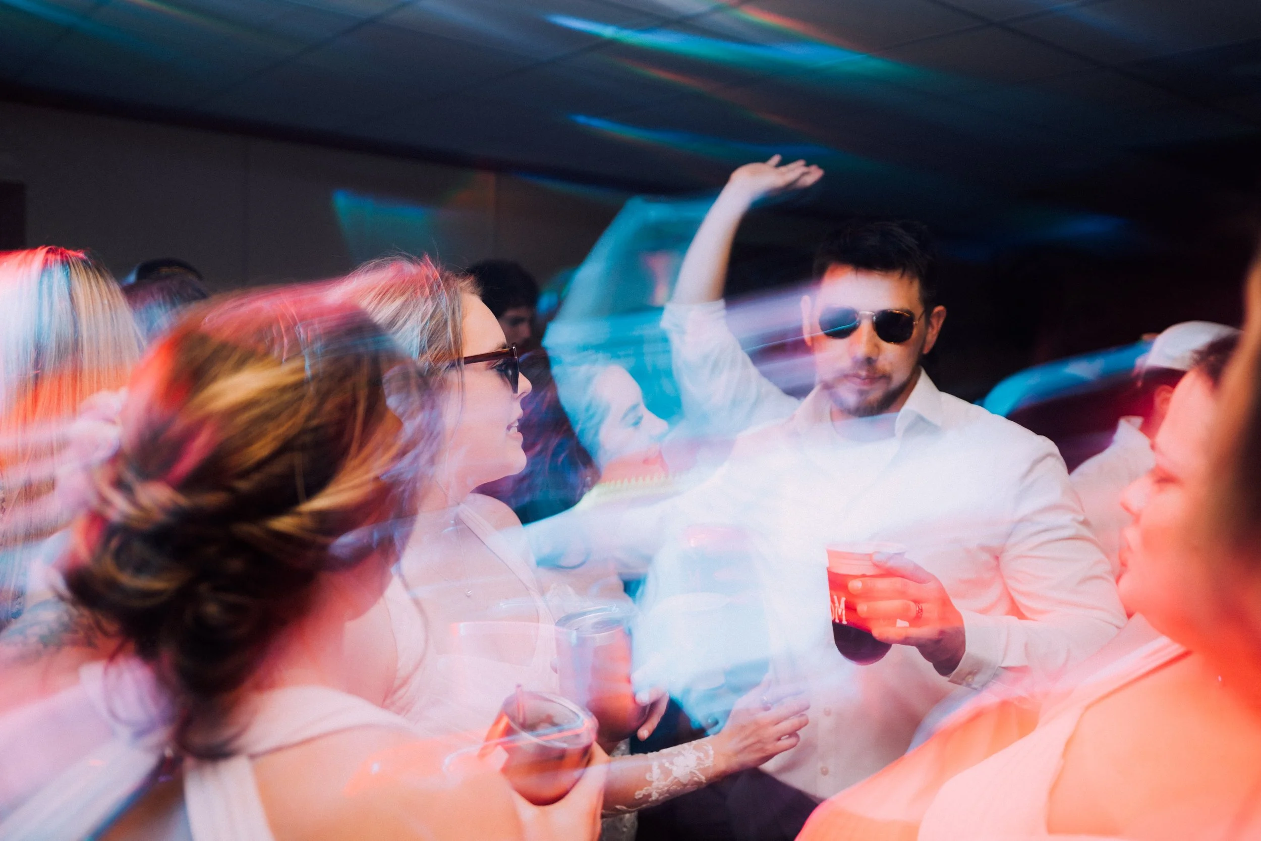Group of young people at a party dancing and socializing, with one guy wearing sunglasses and holding a drink.