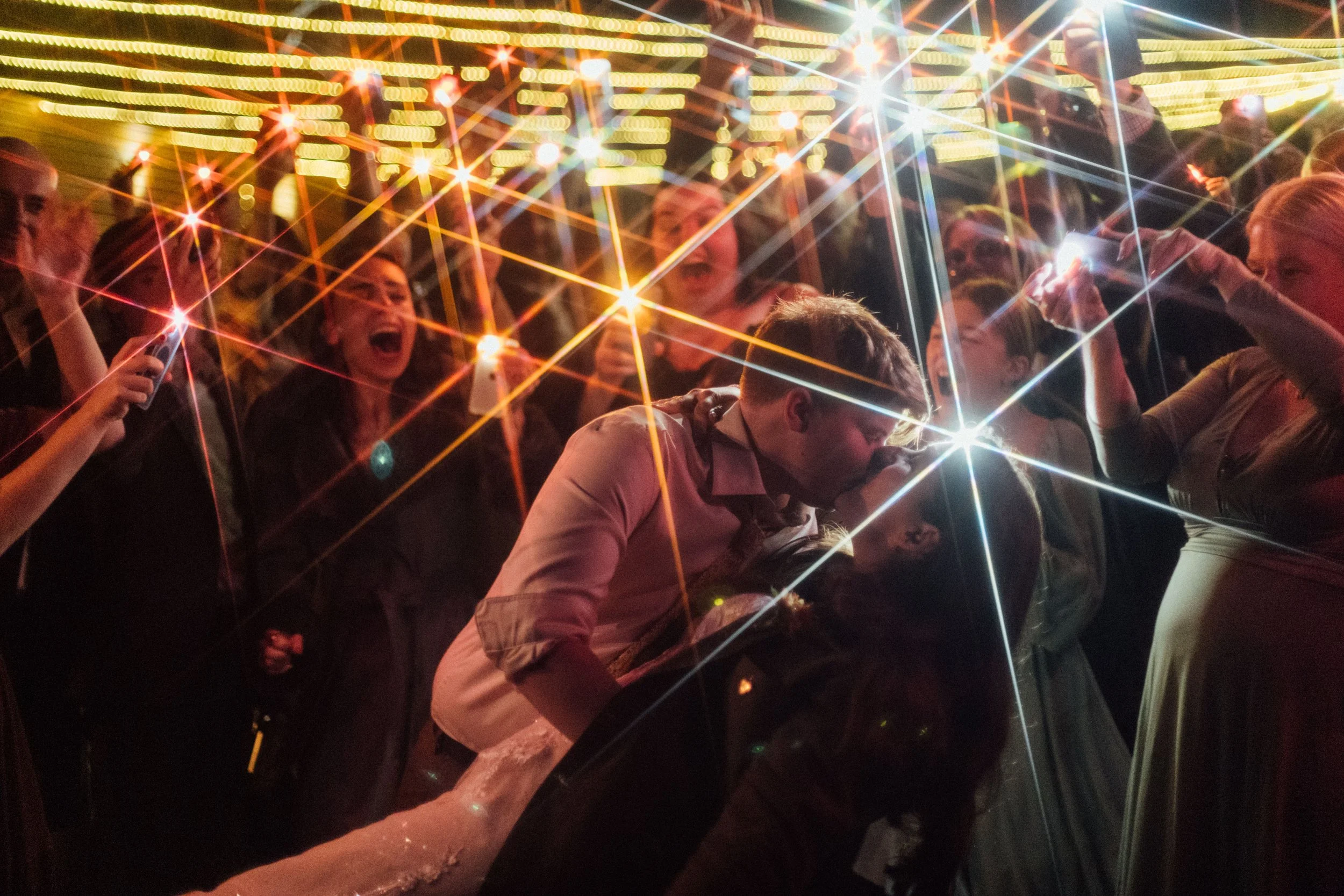 People taking photos and shouting in a lively night setting with bright lights and colorful reflections.