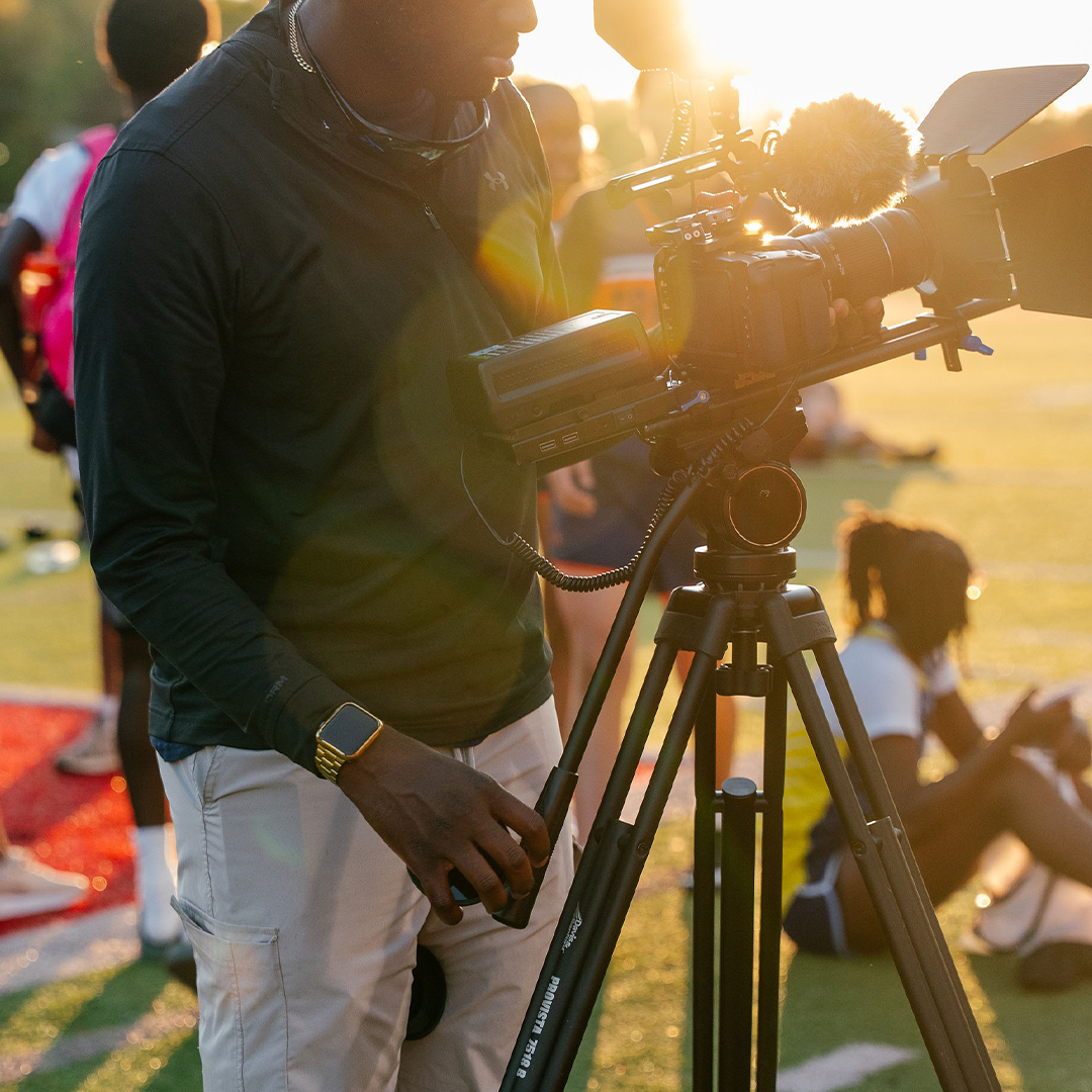 Studio Merv operates a professional video camera on a tripod at an outdoor event, backlit by a golden sunset as people gather nearby.