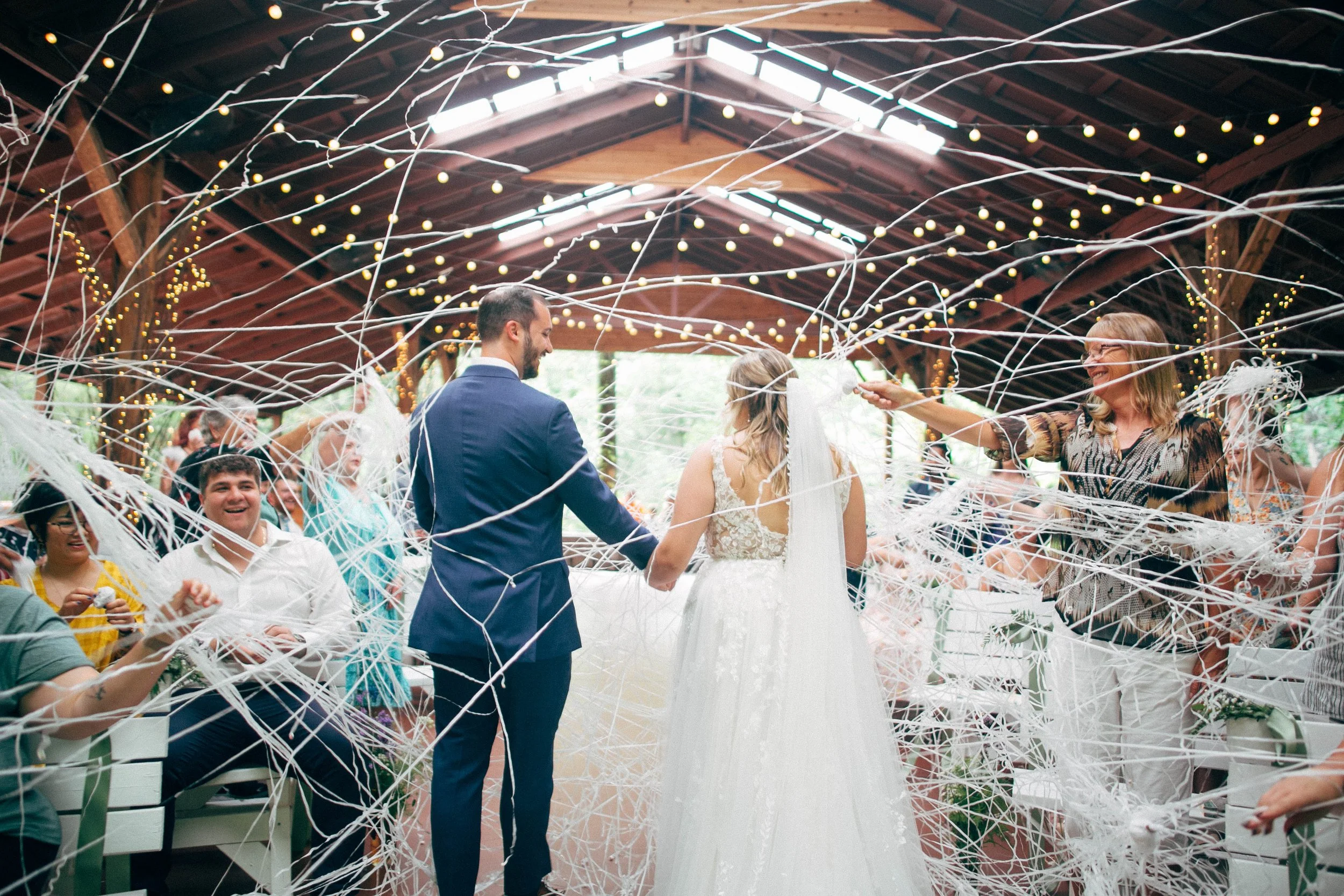 A bride and groom are exchanging vows during a wedding ceremony under a wooden pavilion decorated with string lights, surrounded by guests and white streamers.