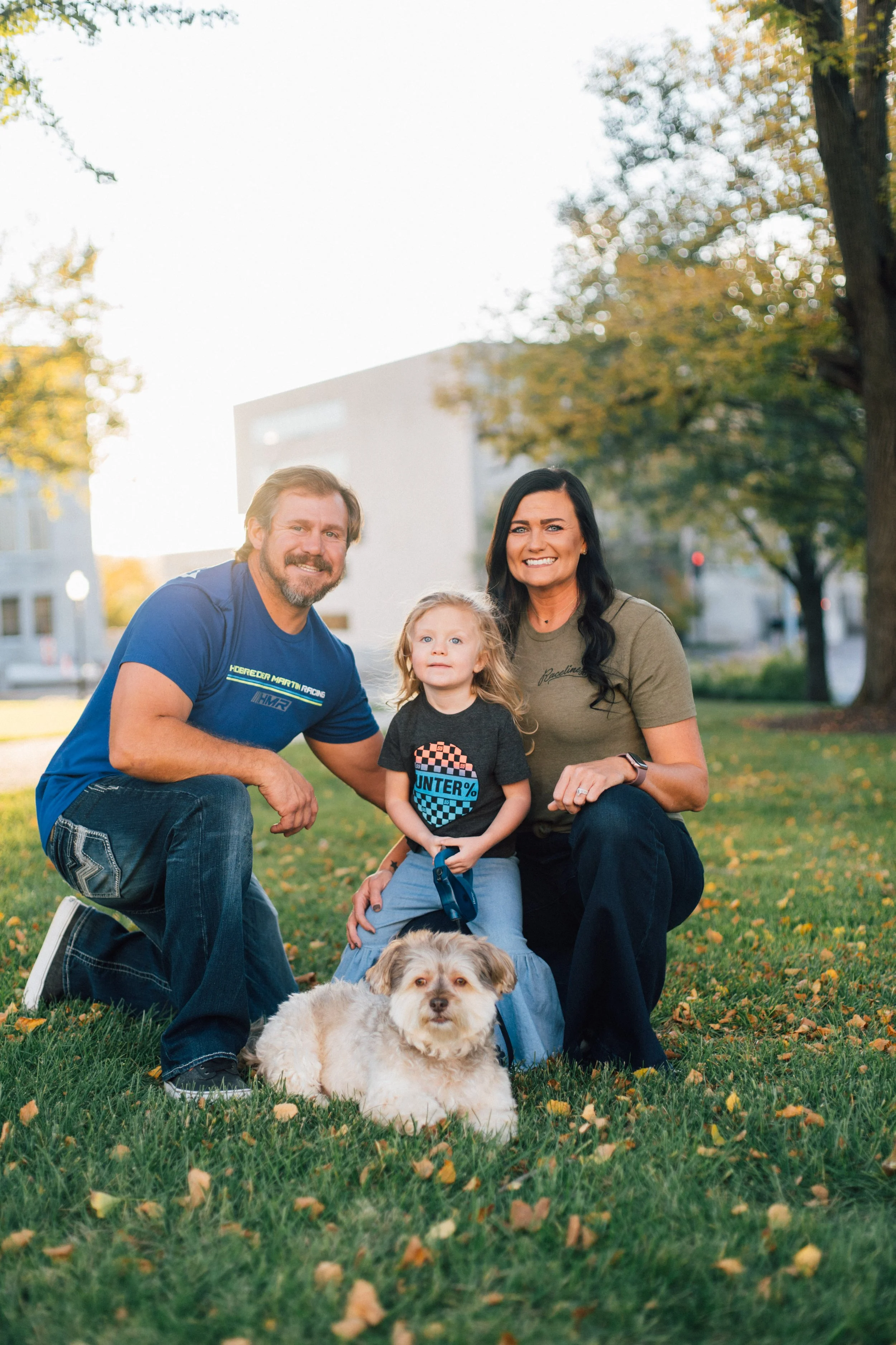 A family of three, including a man, woman, and a young girl, along with their dog, enjoying time together outdoors in a park with autumn leaves on the grass. The man crouches on the left, the woman and girl are kneeling, and the dog is sitting in front of them.