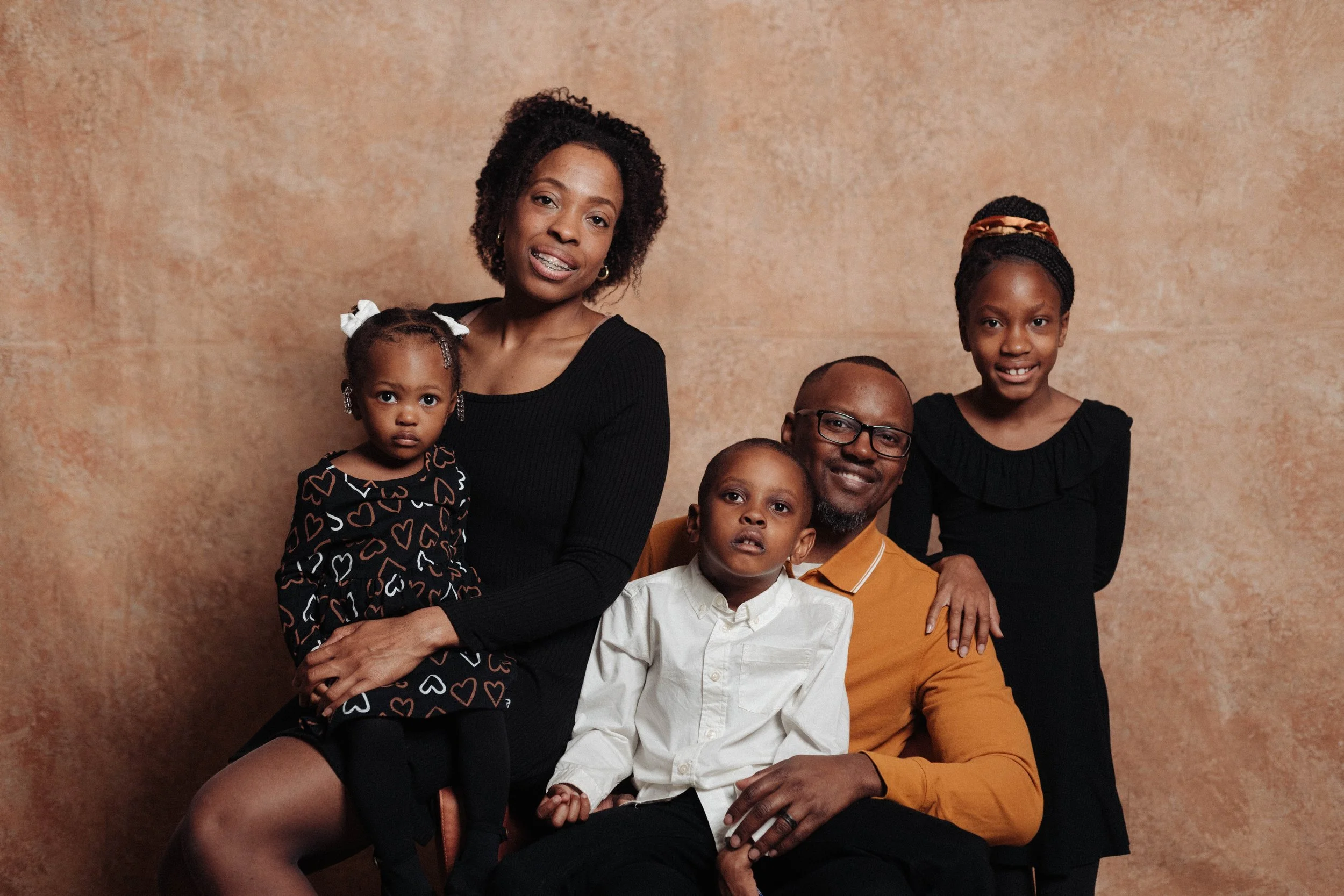 A multi-generational Black family portrait with four children, a young woman, and a man all smiling and posing against a brown textured background.