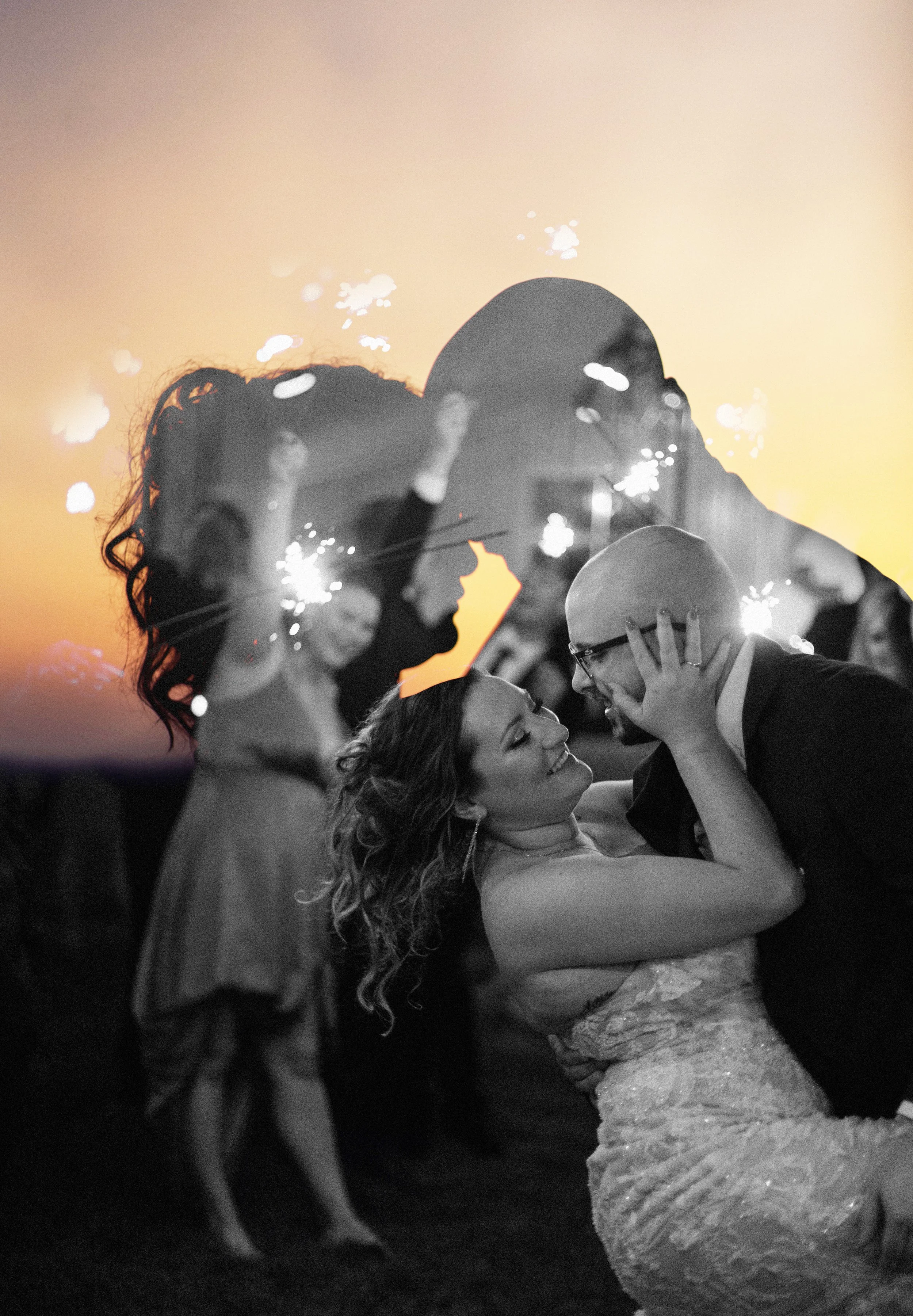 Couple dancing at a wedding reception with sparklers, one woman in a wedding dress and a man in a tuxedo, smiling and embracing each other.