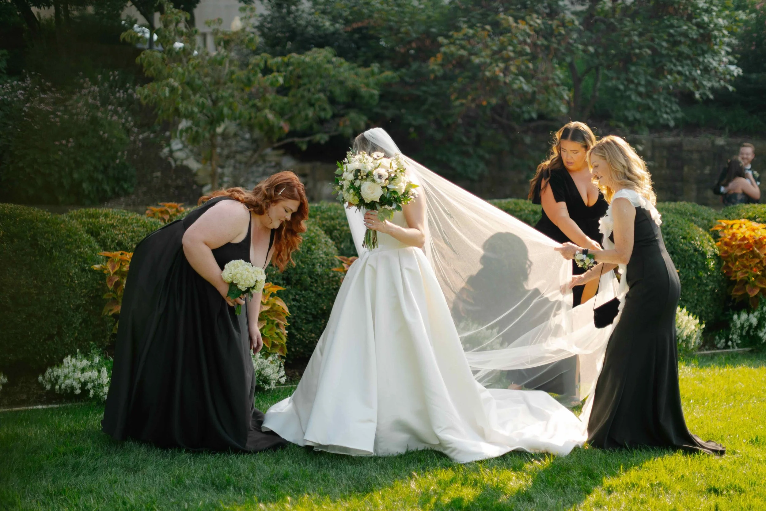 A bride in a white wedding gown holding a bouquet, surrounded by three women in black dresses, as they prepare the train of her dress outdoors in a garden with trees and bushes.