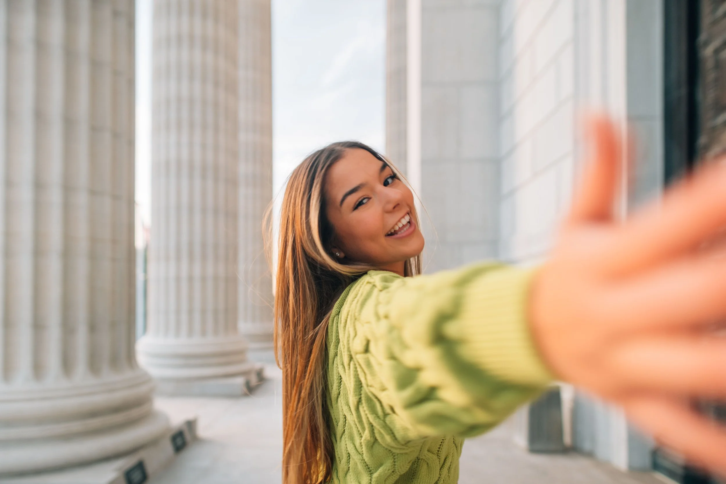 A young woman with long hair smiling and reaching out with her right hand towards camera, standing in front of white columns, wearing a bright green sweater in an outdoor setting with large classical columns.