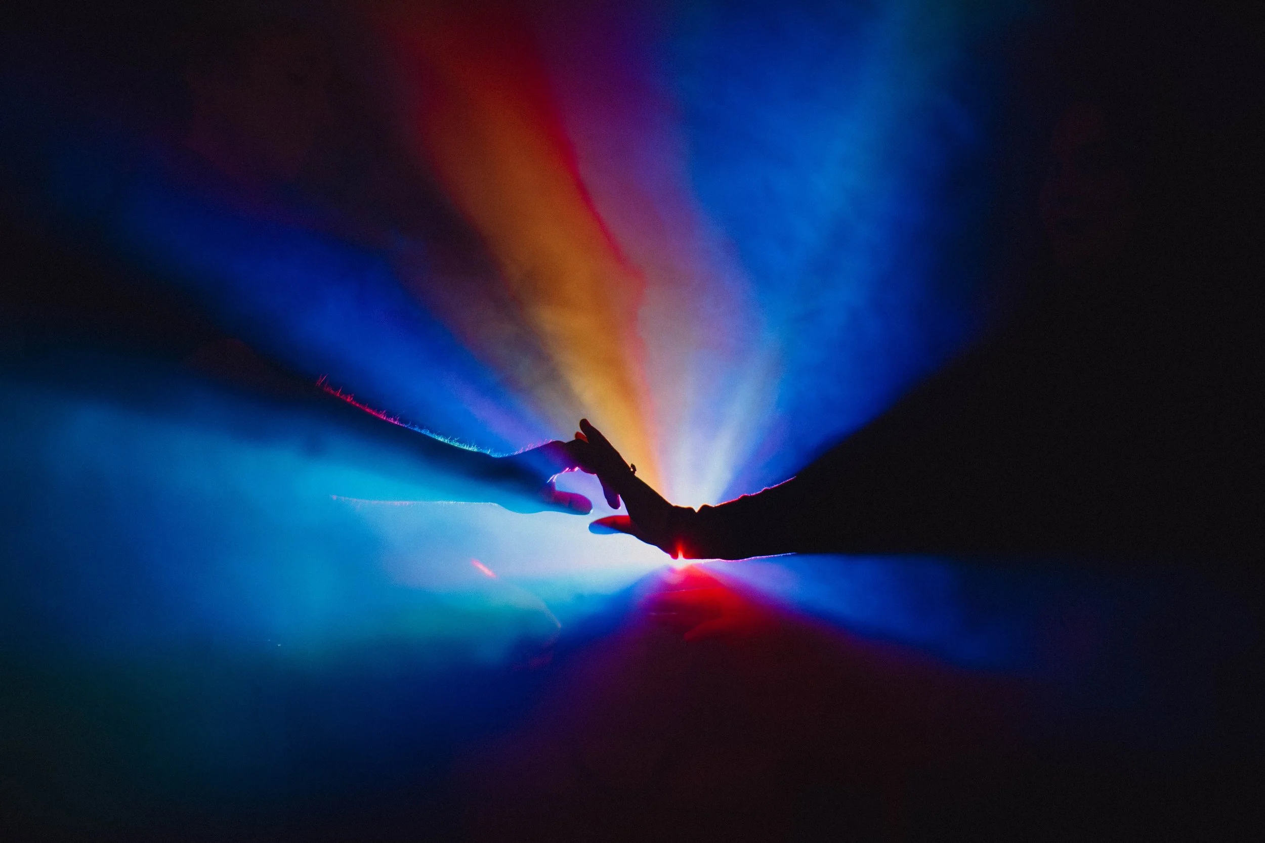 Two hands reaching toward each other with a colorful, luminous backdrop of red, blue, and yellow light beams