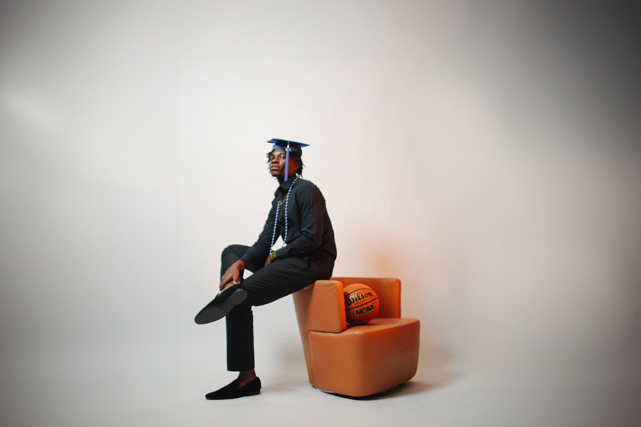 A young man wearing a graduation cap and sitting on a chair with one leg crossed over the other, positioned on top of a basketball. The scene is minimalistic with a plain, light-colored background.