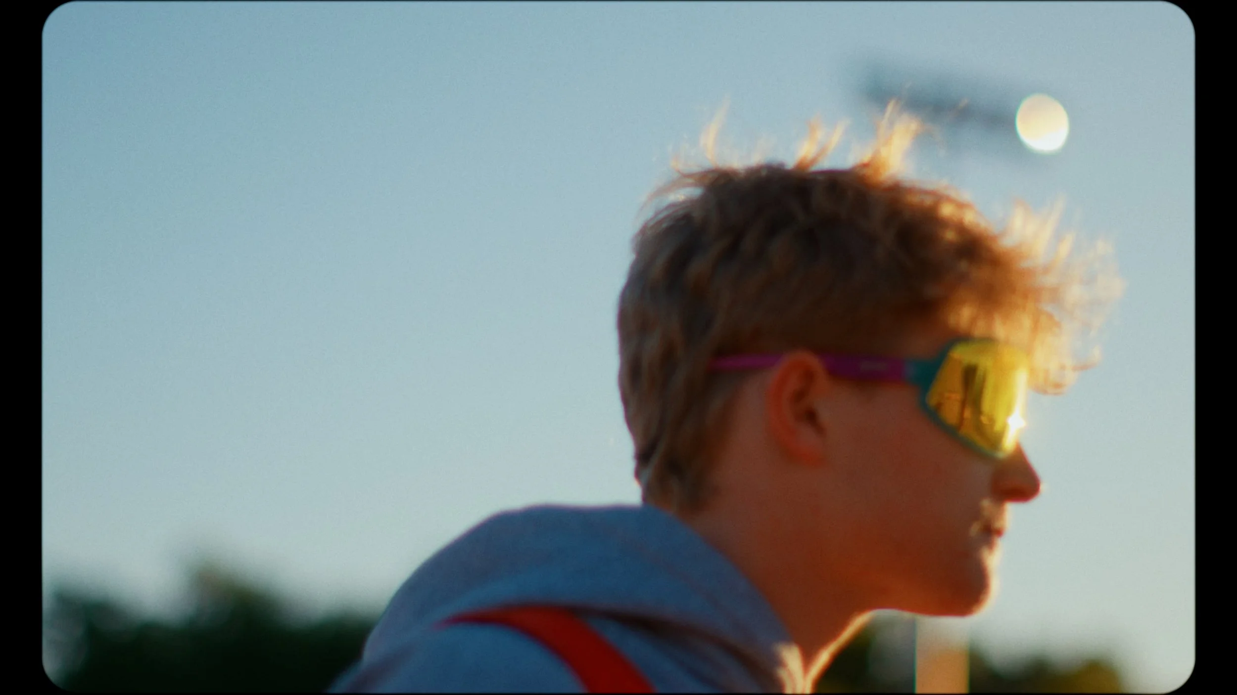 A young boy with messy, sunlit hair wearing yellow sunglasses and a blue jacket with red trim, outdoors during sunset or sunrise.