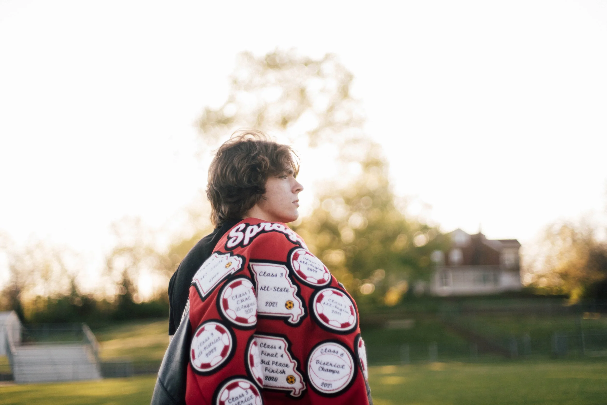 Profile of a young man with curly brown hair standing outdoors during sunset, wearing a letterman jacket with various sports patches, in front of trees and a residential area.