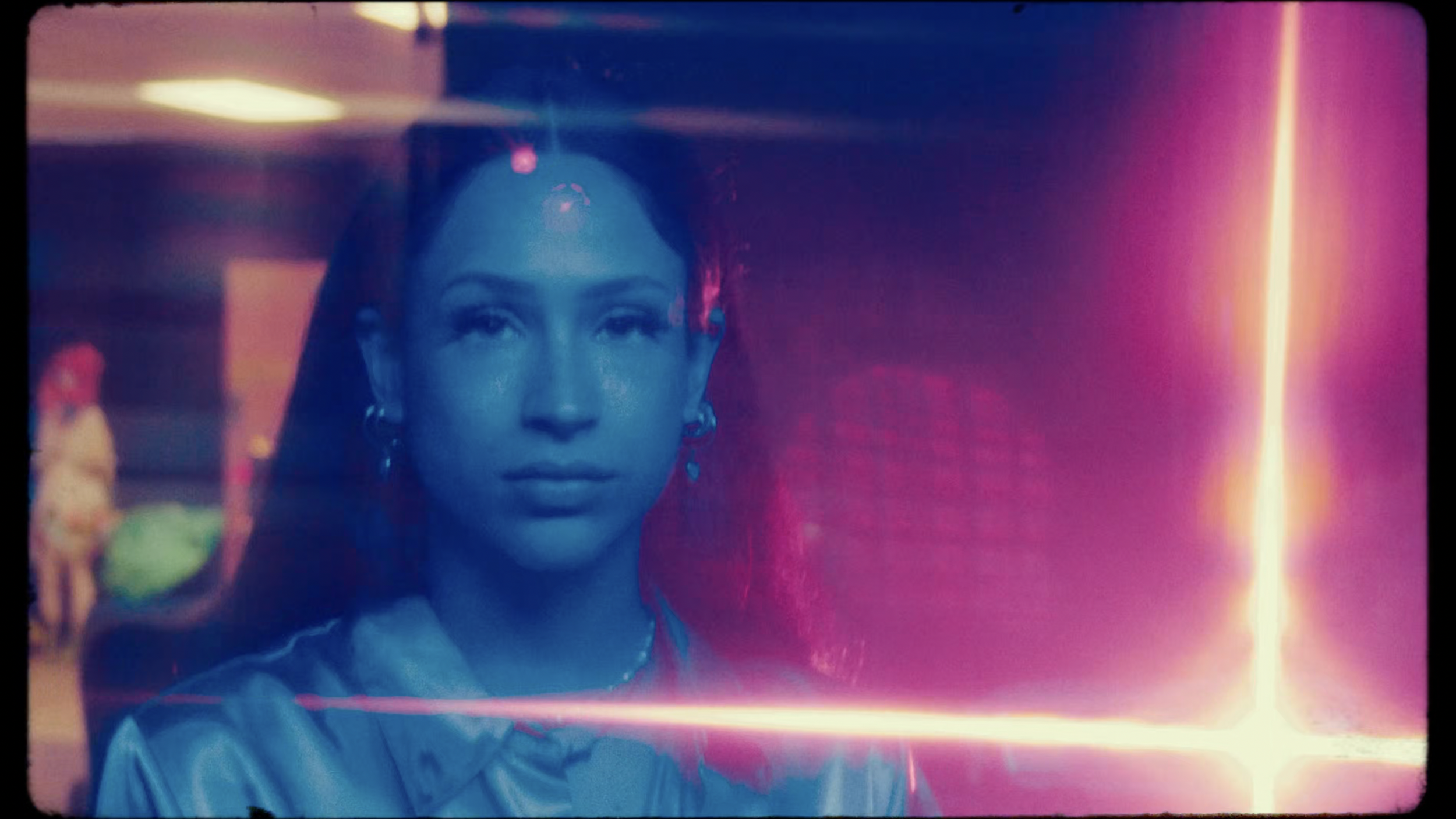 A woman with earrings looking directly at the camera, illuminated by colorful neon lights in a vibrant, artistic setting.
