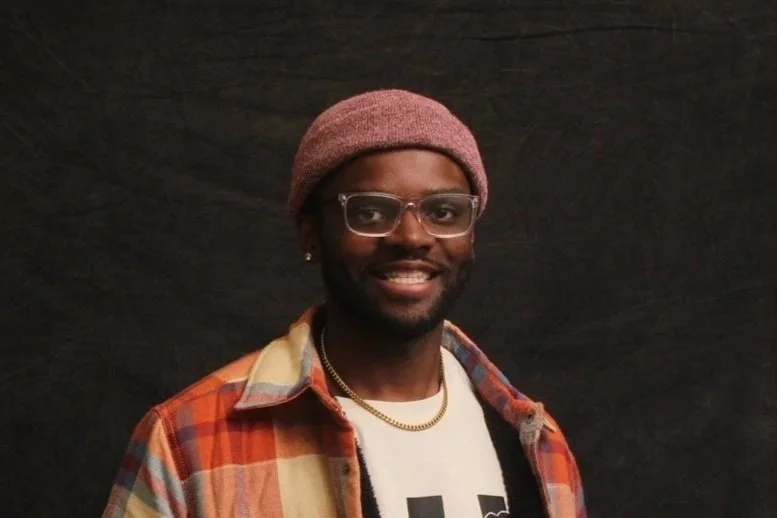 A smiling man with glasses, a pink knit hat, a gold chain, and a plaid shirt over a white graphic tee against a black background.