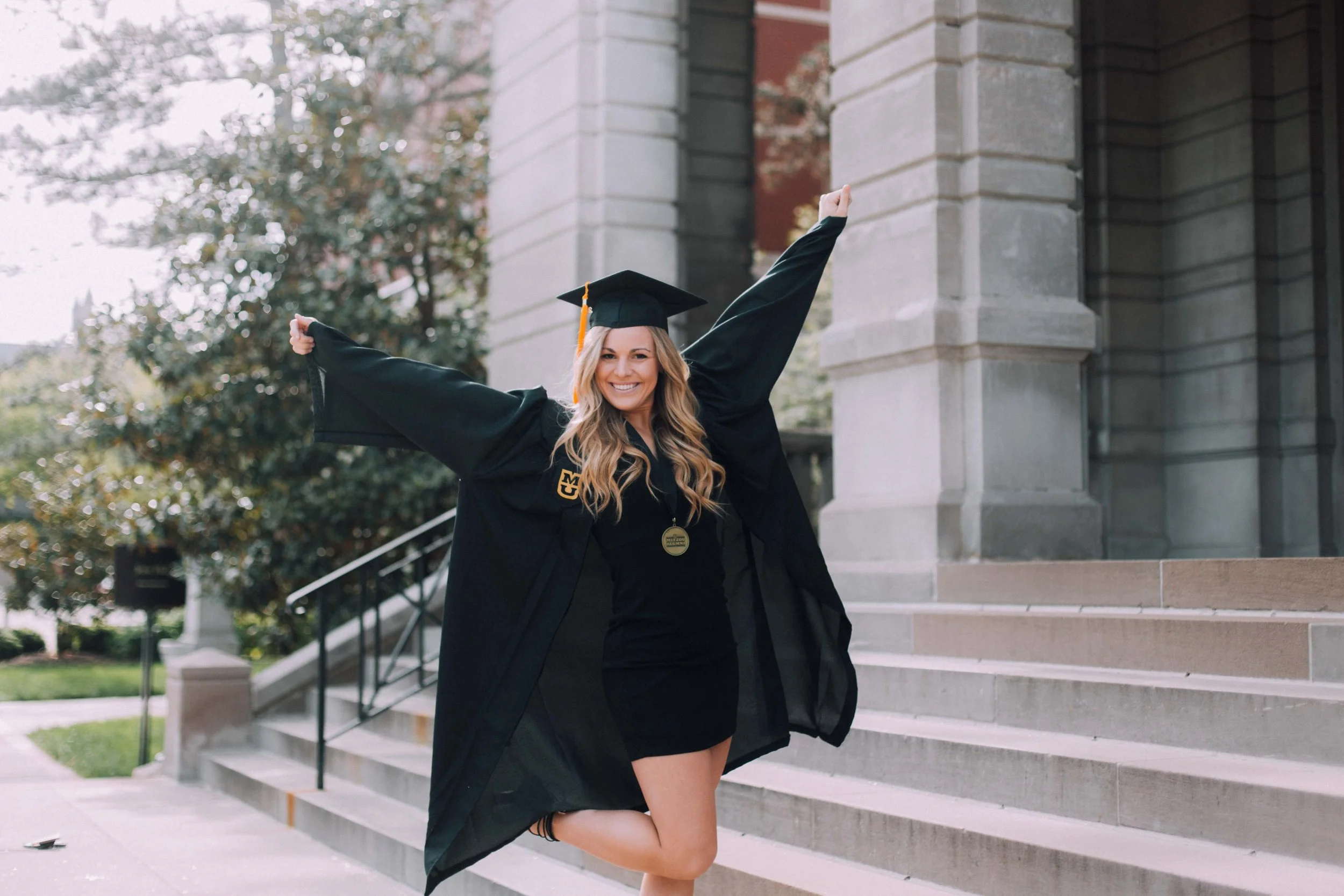 A woman in a black graduation gown and cap celebrating with arms raised on steps outside of a building.